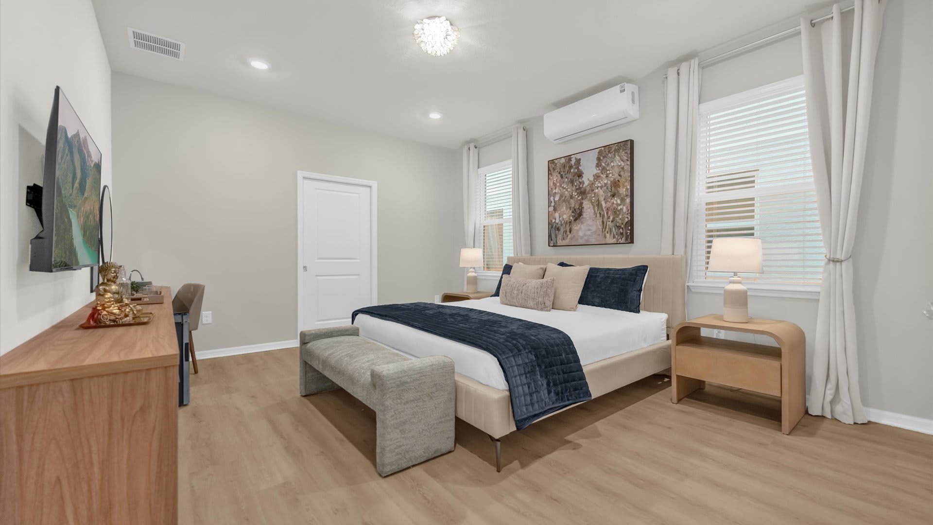 A bright bedroom with light wood-look flooring, a queen-sized bed with neutral bedding, and two large windows with white blinds. A wall-mounted AC unit and a small desk with a TV are visible.