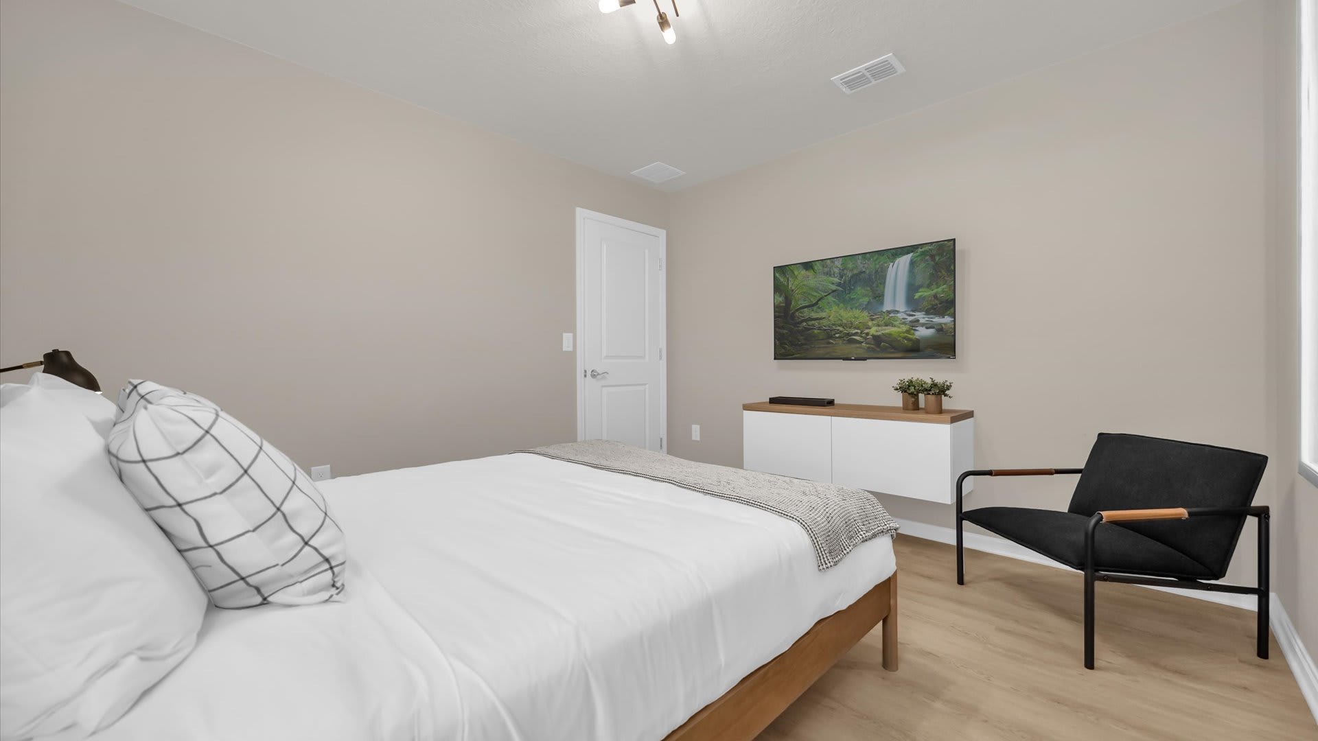 Another angle of the bedroom, showcasing a wall-mounted TV above a floating white and wood console. A modern black armchair sits in the corner, with light wood flooring extending throughout the room.