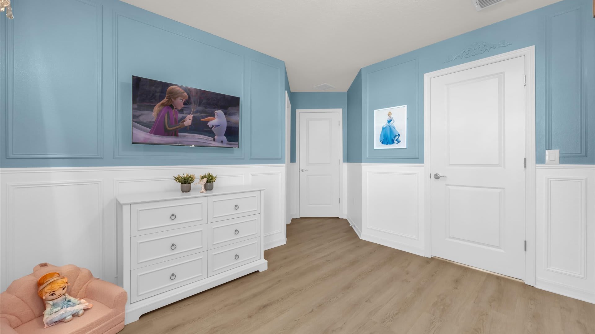 Princess-themed bedroom with light blue paneled walls and white wainscoting. A wall-mounted TV, a white dresser, and a small pink armchair are visible, along with white doors.