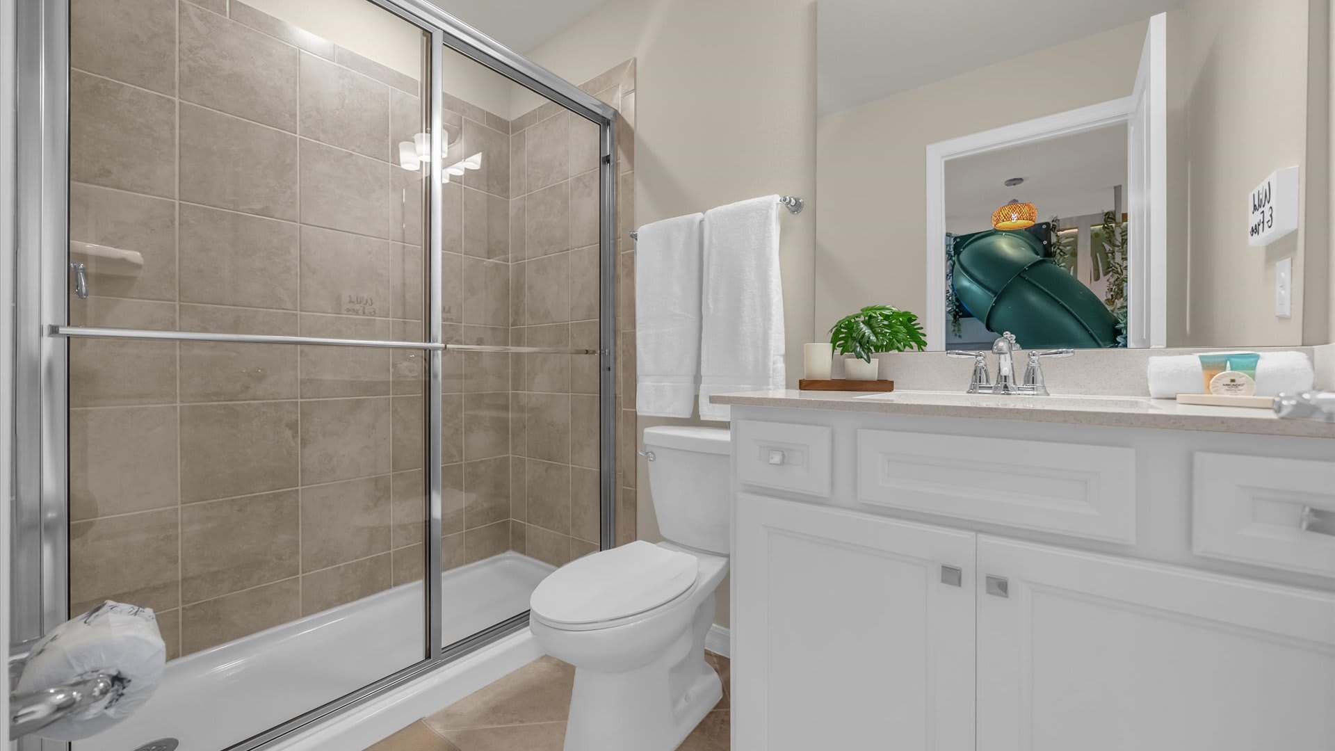 Modern bathroom featuring a glass-enclosed shower with beige tiled walls and a white toilet. A single vanity with a light countertop and white cabinets is visible, along with a mirror reflecting the themed bedroom.