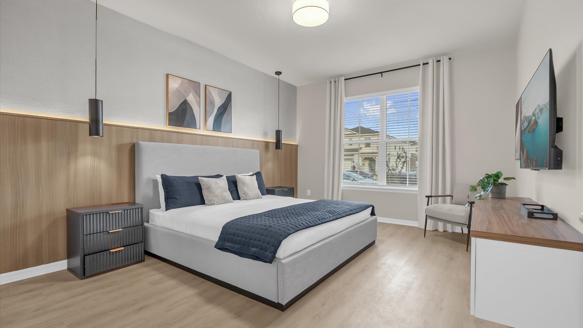 Contemporary bedroom featuring a light gray upholstered king bed with dark blue bedding, a wood-paneled accent wall with integrated lighting, and two dark pendant lights. A large window with blinds and curtains overlooks a neighborhood.
