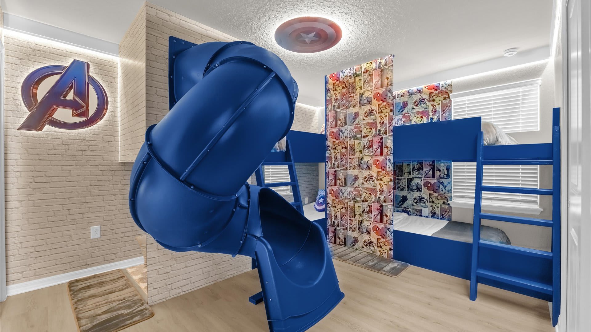 A vibrant, themed kids' bedroom featuring a blue bunk bed, a large blue slide, and a Captain America shield light fixture on the ceiling. The walls are adorned with a light brick pattern and comic book wallpaper.