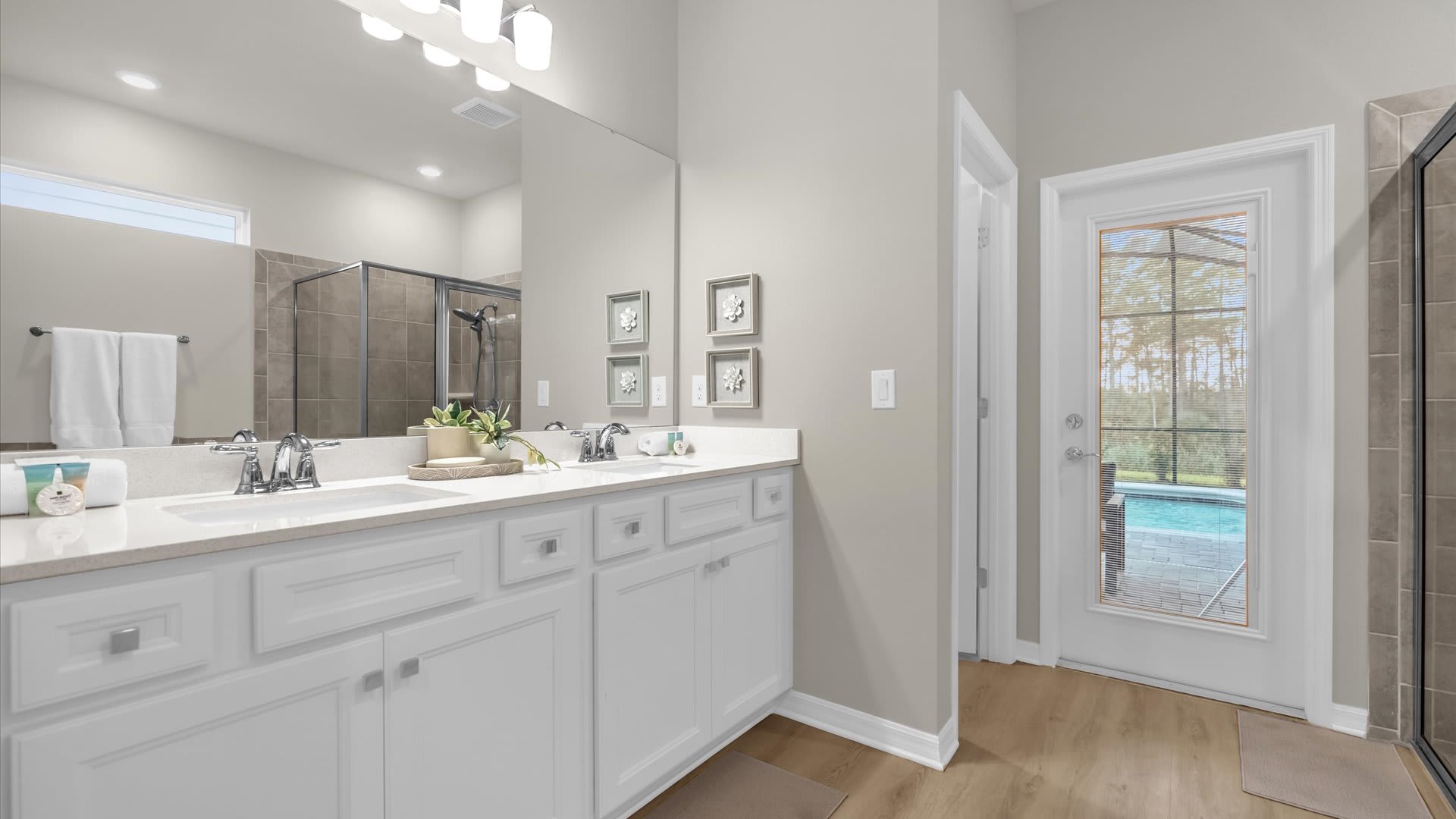 A spacious bathroom featuring a double white vanity with light countertops and a large mirror. A door with a view of an outdoor pool area is visible, and a glass-enclosed shower is partially seen.
