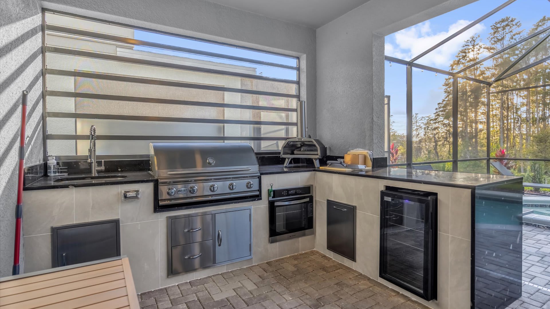 A well-equipped outdoor kitchen on a screened patio features a built-in grill, oven, sink, and beverage cooler, with black granite countertops and a view of the pool area and natural landscape.