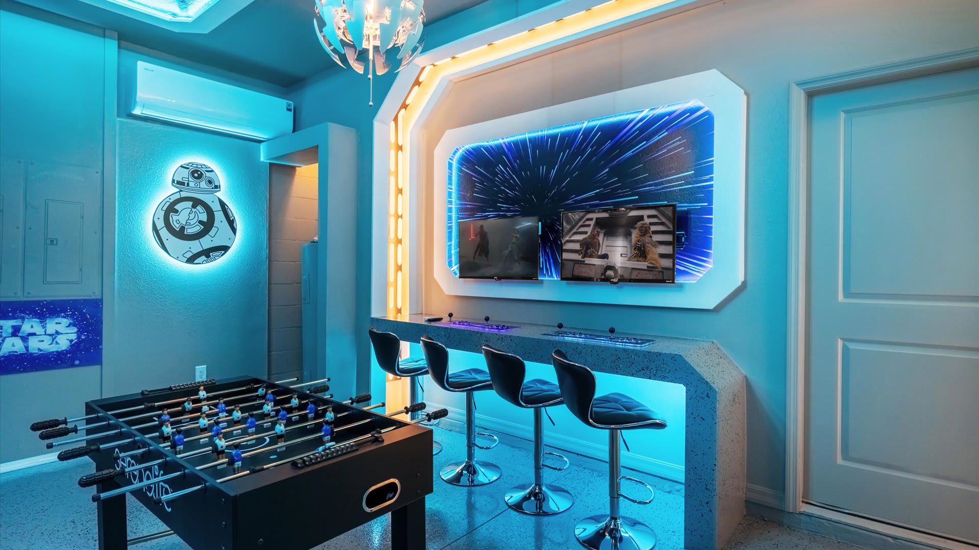 Themed game room with blue LED lighting, featuring a foosball table and a gaming station with multiple screens and bar stools. A large BB-8 Star Wars character cutout with blue backlighting adorns the wall.