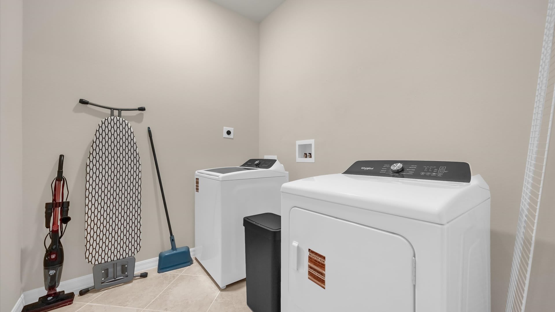 Functional laundry room equipped with a white top-load washing machine and a white dryer. An ironing board, broom, and vacuum cleaner are stored neatly against the light beige wall.