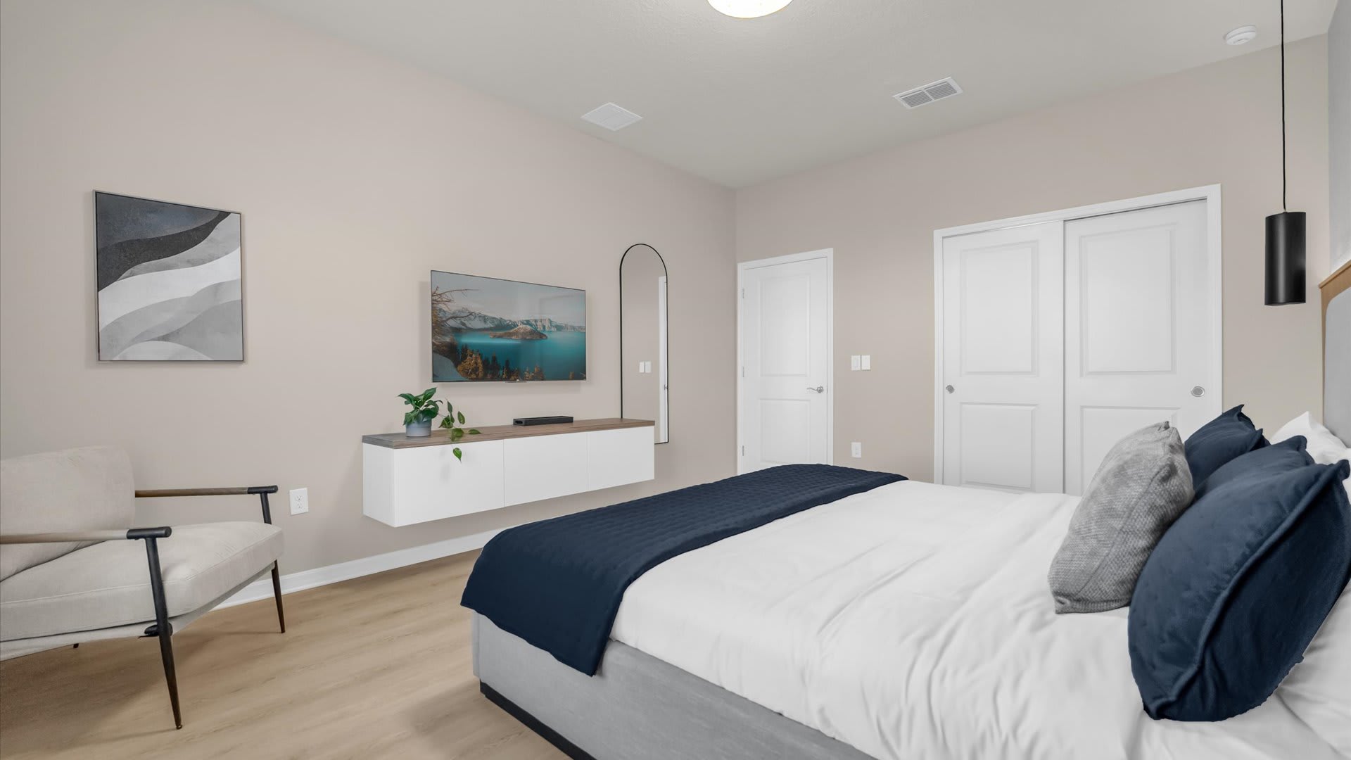 View of a modern bedroom with a light gray upholstered bed, dark blue bedding, and light wood-look flooring. A wall-mounted TV and a floating console are visible, along with a full-length mirror and two white closet doors.