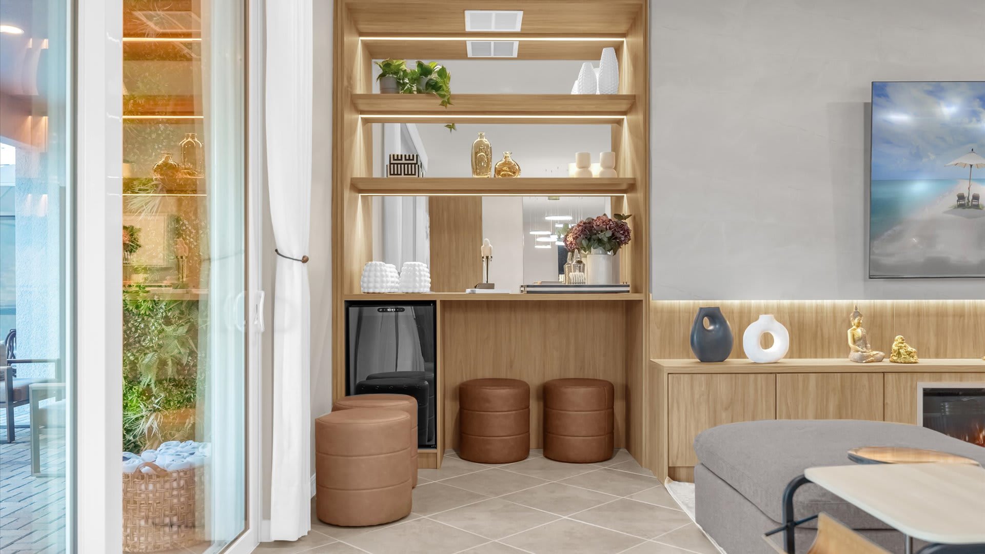 Detail shot of a built-in light wood shelving unit in the living room, featuring mirrored back panels and accent lighting. Decorative vases, plants, and a mini-fridge are visible, with two brown leather ottomans below, and sliding glass doors to the left.