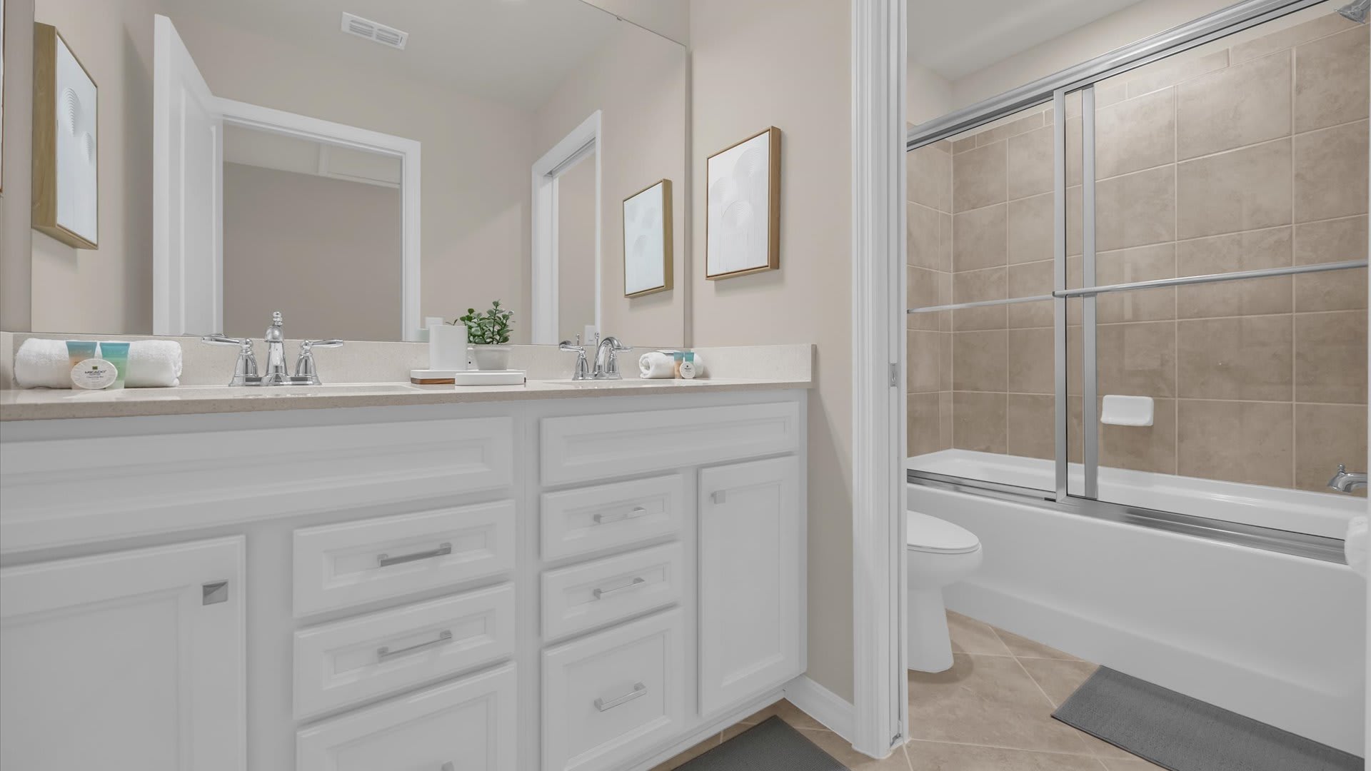 Modern bathroom with a double white vanity, light beige walls, and a shower/tub combo with a sliding glass door and beige tiled walls. Two framed pictures hang above the vanity.