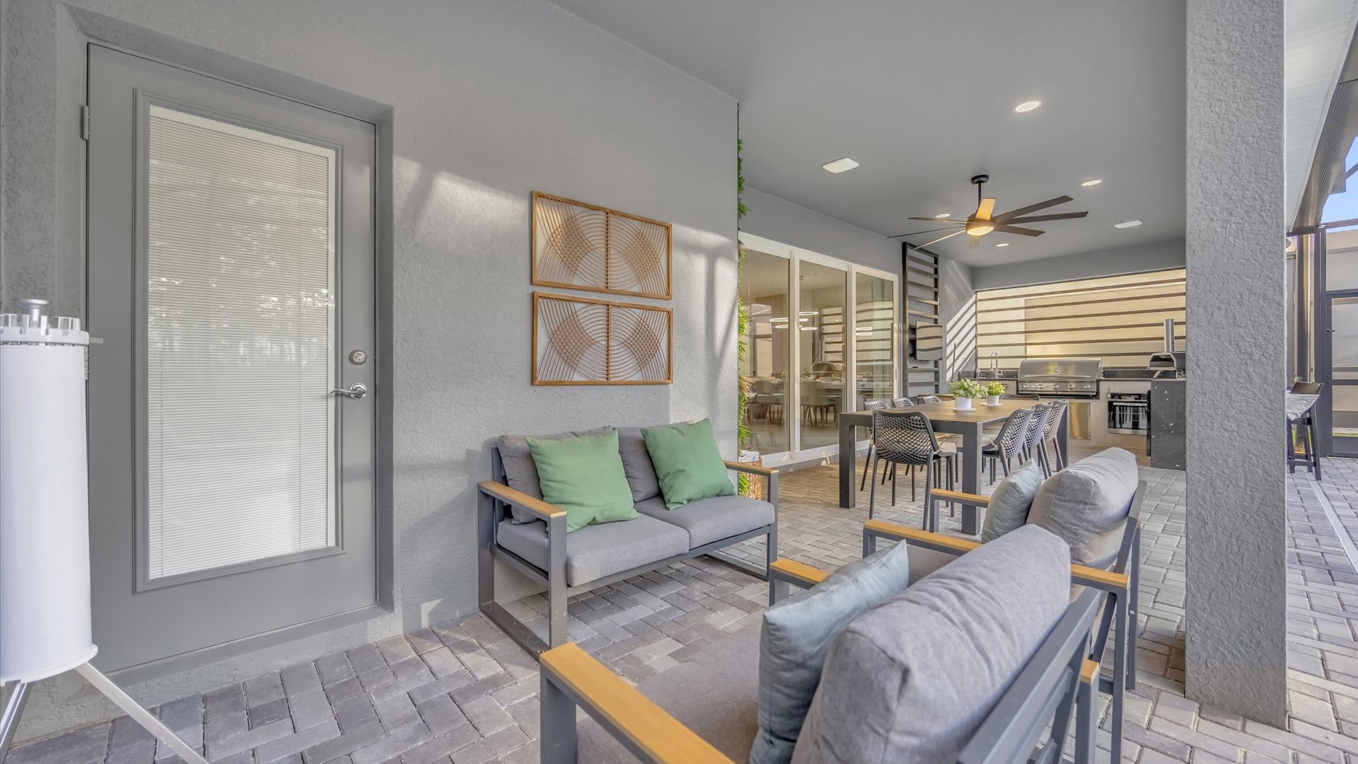 A covered outdoor lounge area on a paved patio features comfortable grey sofas with green and blue cushions, offering a relaxed space adjacent to the main outdoor dining and kitchen area.