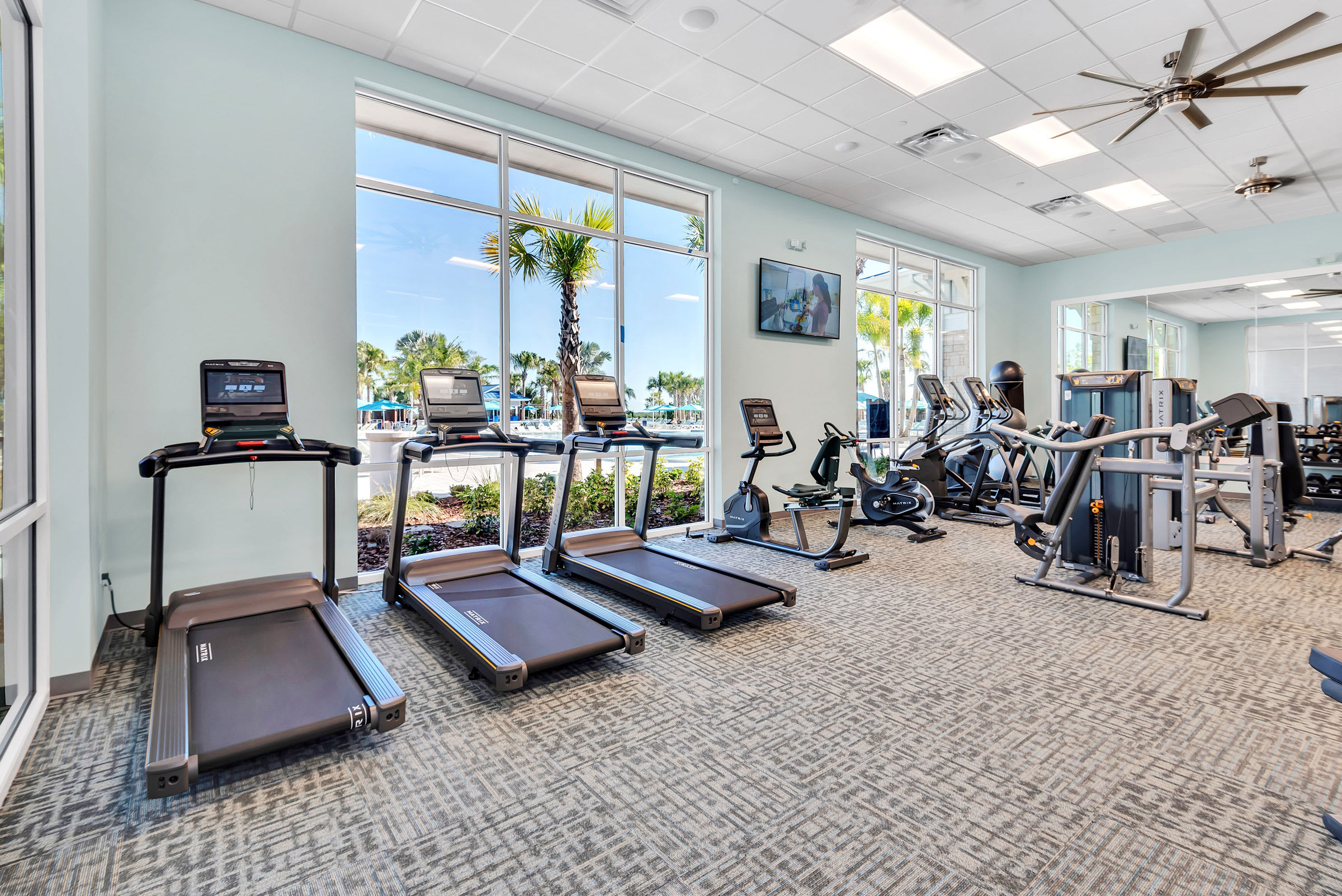 Bright fitness center with several treadmills and exercise bikes facing large windows that offer views of palm trees and the resort's outdoor amenities, featuring light-colored walls and patterned carpet.