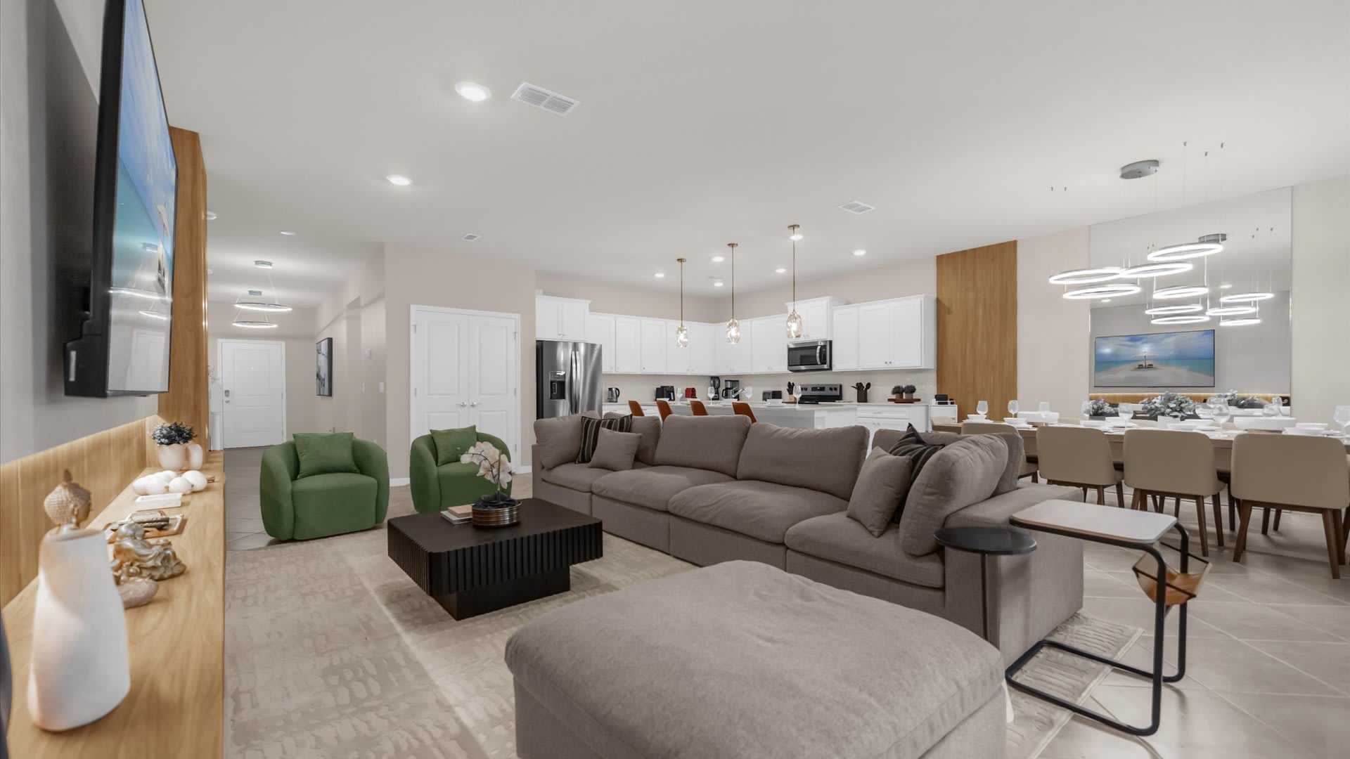 An open-concept living room features a large L-shaped sectional sofa, a dark coffee table, and two green armchairs, flowing into a modern kitchen and dining area with contemporary lighting.