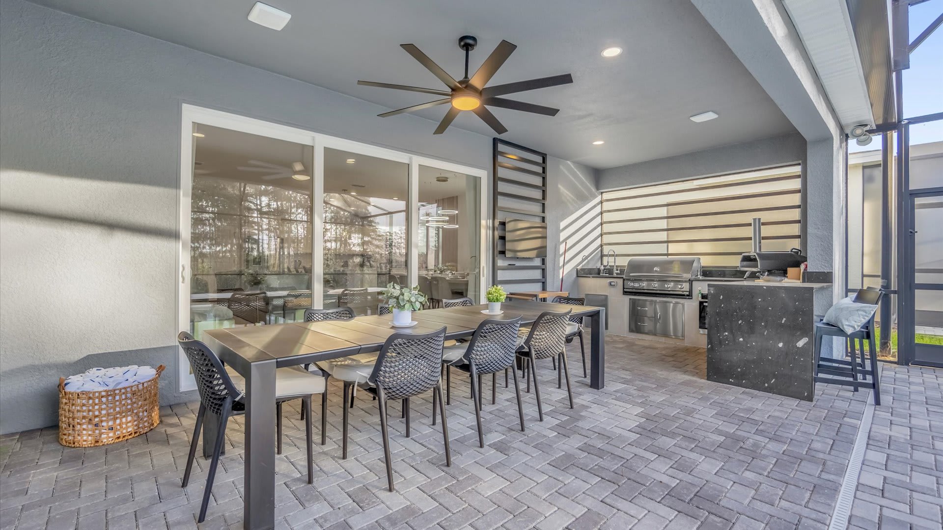 A spacious covered outdoor dining area on a paved patio features a large table with seating for eight, a ceiling fan, and a view into the indoor dining room through sliding glass doors.