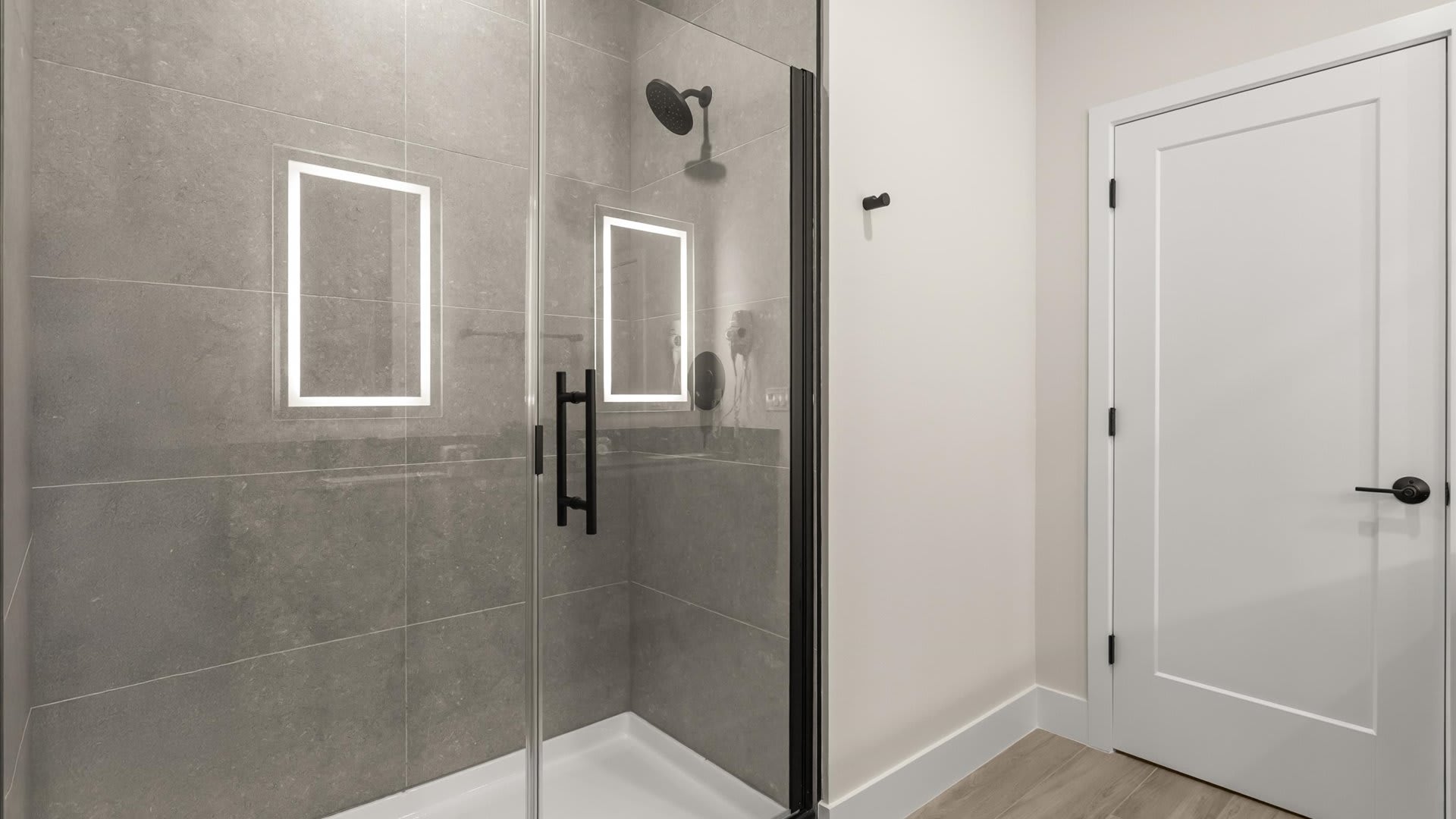 Contemporary walk-in shower with grey large-format tiled walls, a clear glass enclosure with a black frame, and two recessed niches with integrated lighting. A black rain shower head is mounted on the wall.