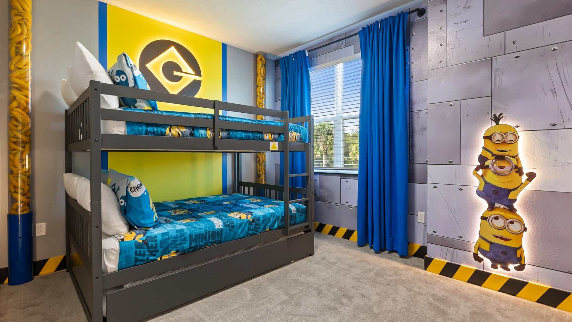Minions-themed kids' bedroom with a grey bunk bed, blue and yellow bedding, and a window with blue curtains. The accent wall features a large yellow 'G' logo, while other walls have industrial-style panels and a glowing Minions character cutout.