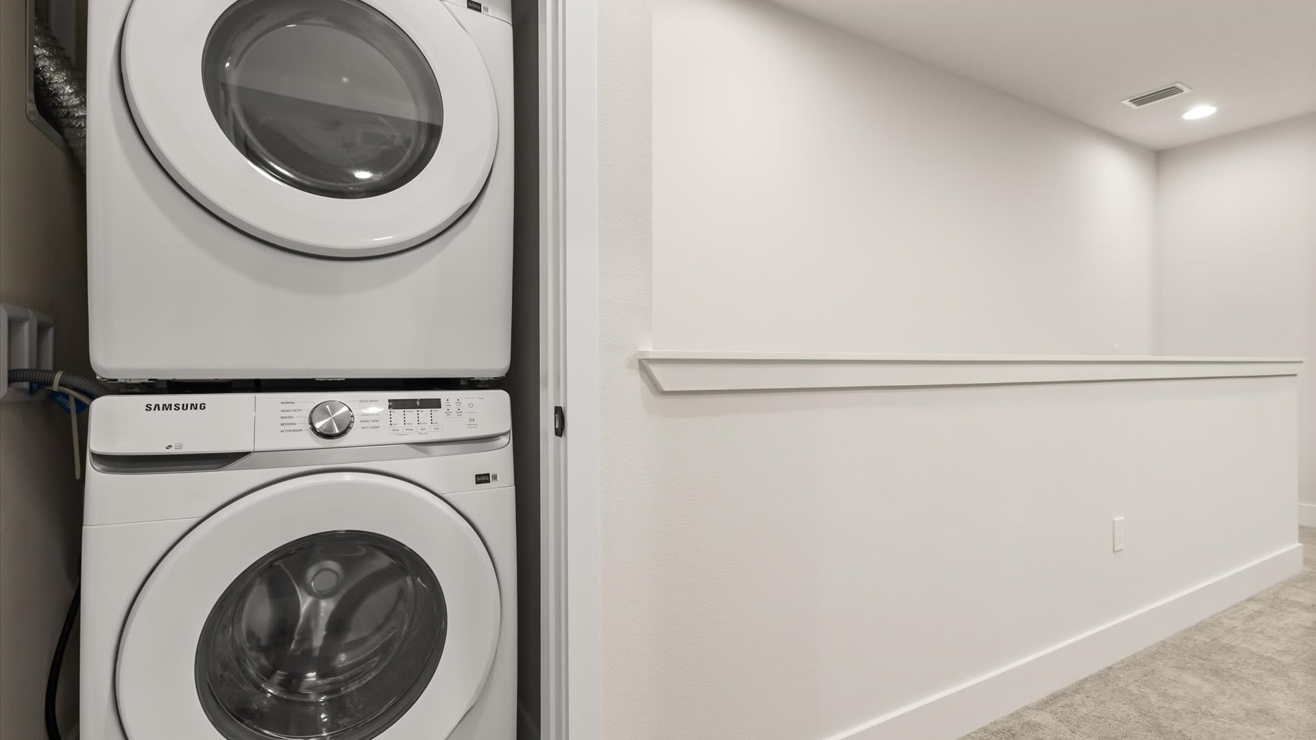 Compact laundry area featuring a stacked white Samsung washer and dryer unit in a closet, with light-colored walls and carpeted flooring. The space is functional and neatly organized.