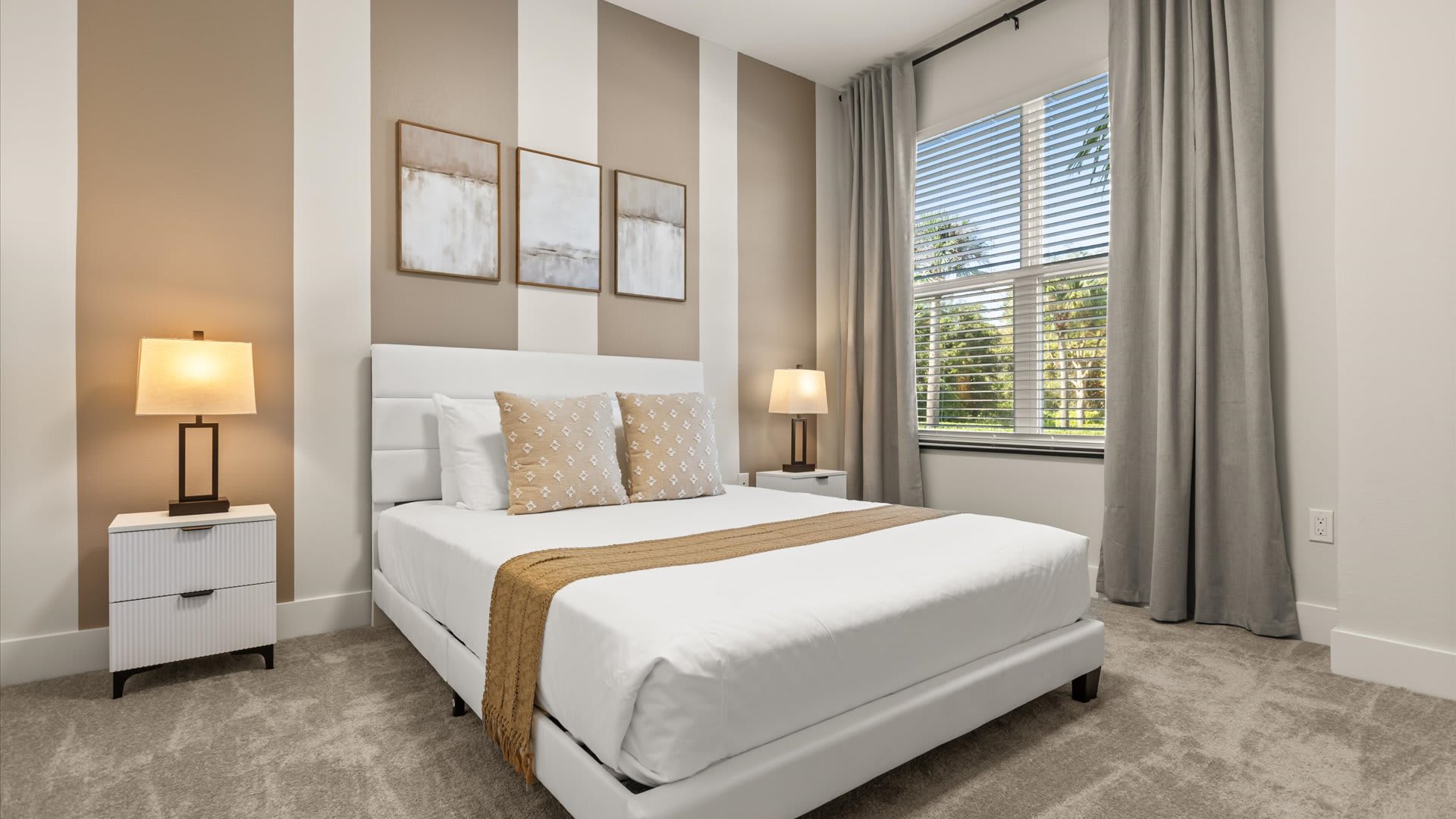 Bright bedroom featuring a white upholstered queen bed with neutral bedding, flanked by two white nightstands with modern lamps. The wall behind the bed has vertical stripes in beige and white, and a large window with blinds and grey curtains offers natural light.