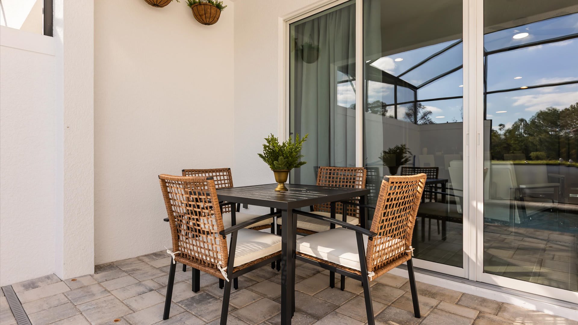 Outdoor dining area on a paved patio, featuring a dark square table with four wicker-style chairs with white cushions, next to large sliding glass doors.