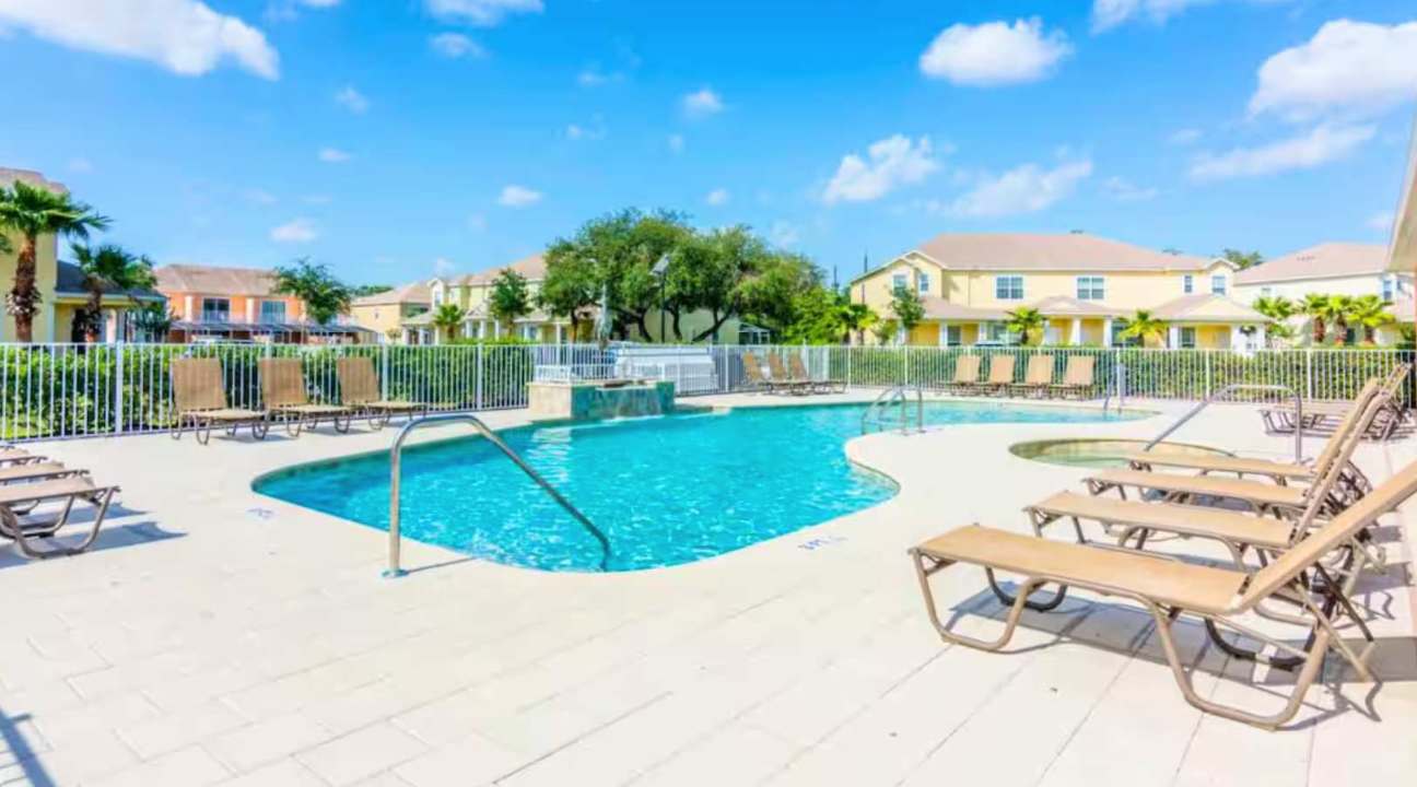 A wide shot of a communal outdoor swimming pool area featuring a freeform pool, a hot tub, and numerous lounge chairs on a paved deck, enclosed by a white fence. Lush green trees and residential buildings are in the background.