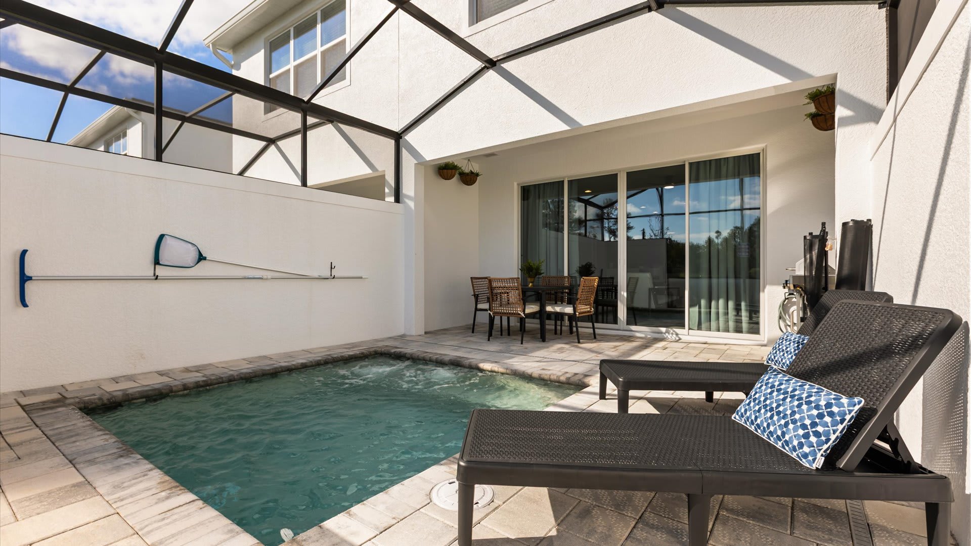 Wide view of the screened outdoor patio with a hot tub, two dark lounge chairs, and an outdoor dining set, showing the sliding glass doors leading into the house.