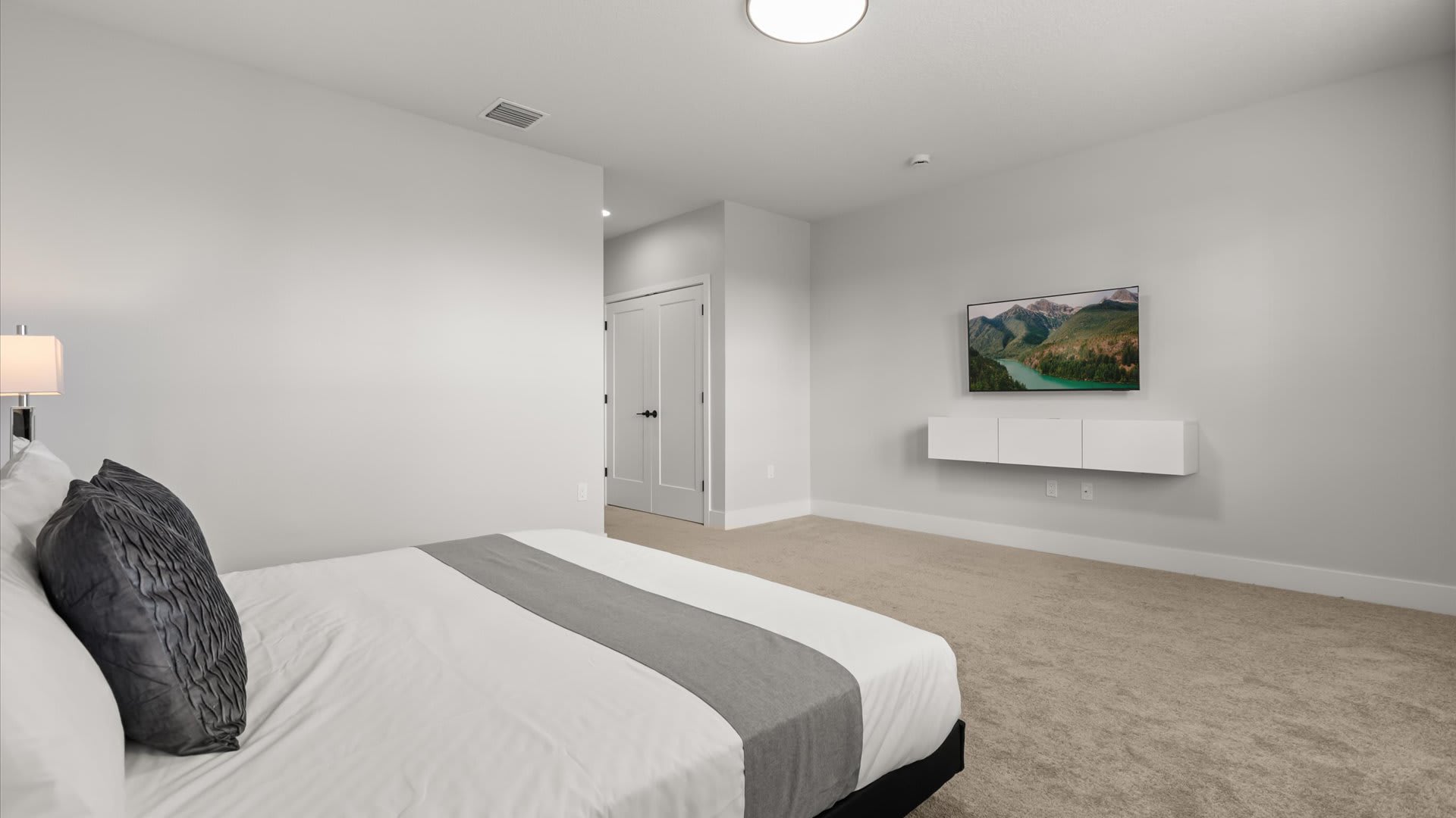 Spacious bedroom with a large bed dressed in white and gray linens, light carpeted flooring, and a wall-mounted TV on a white floating console against a plain white wall.