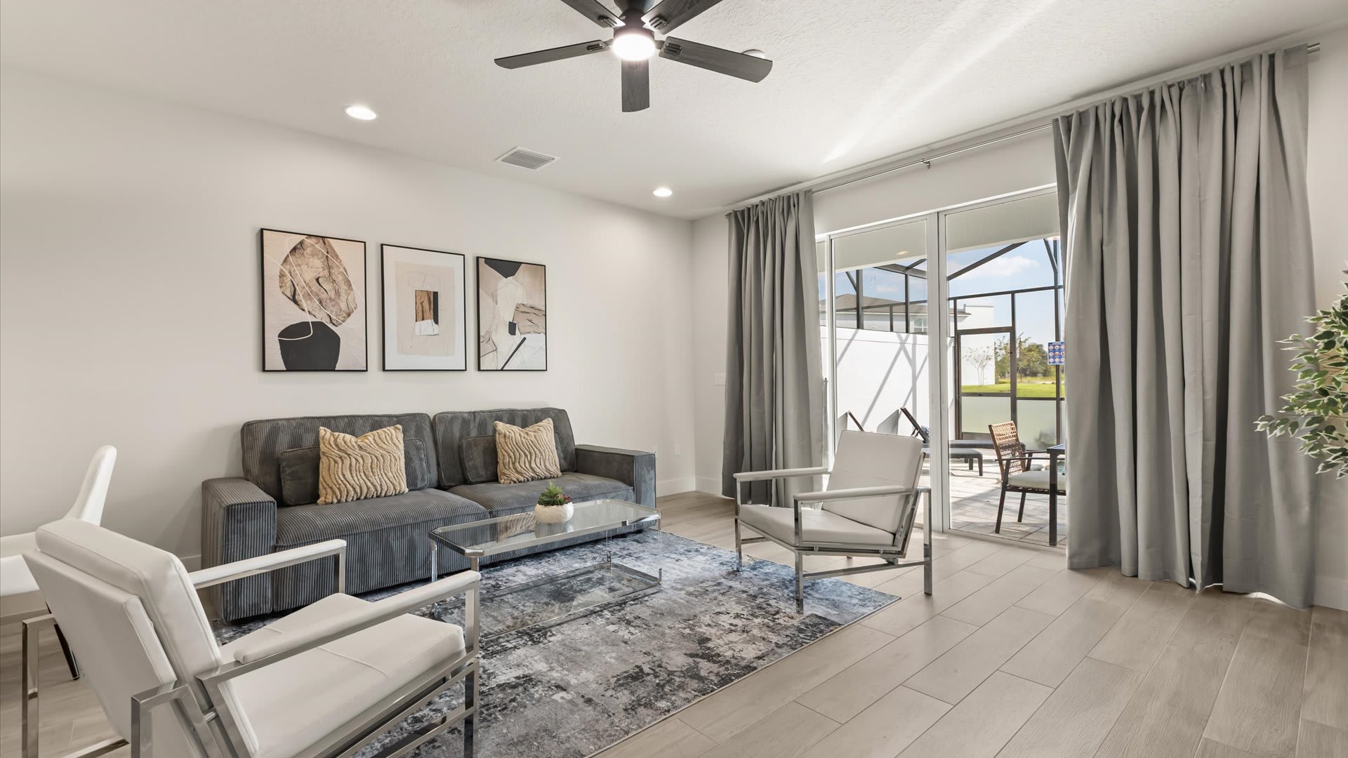 Modern living room featuring a gray sofa with accent pillows, two white armchairs, a glass coffee table on a patterned rug, and sliding glass doors opening to a screened patio with outdoor seating.