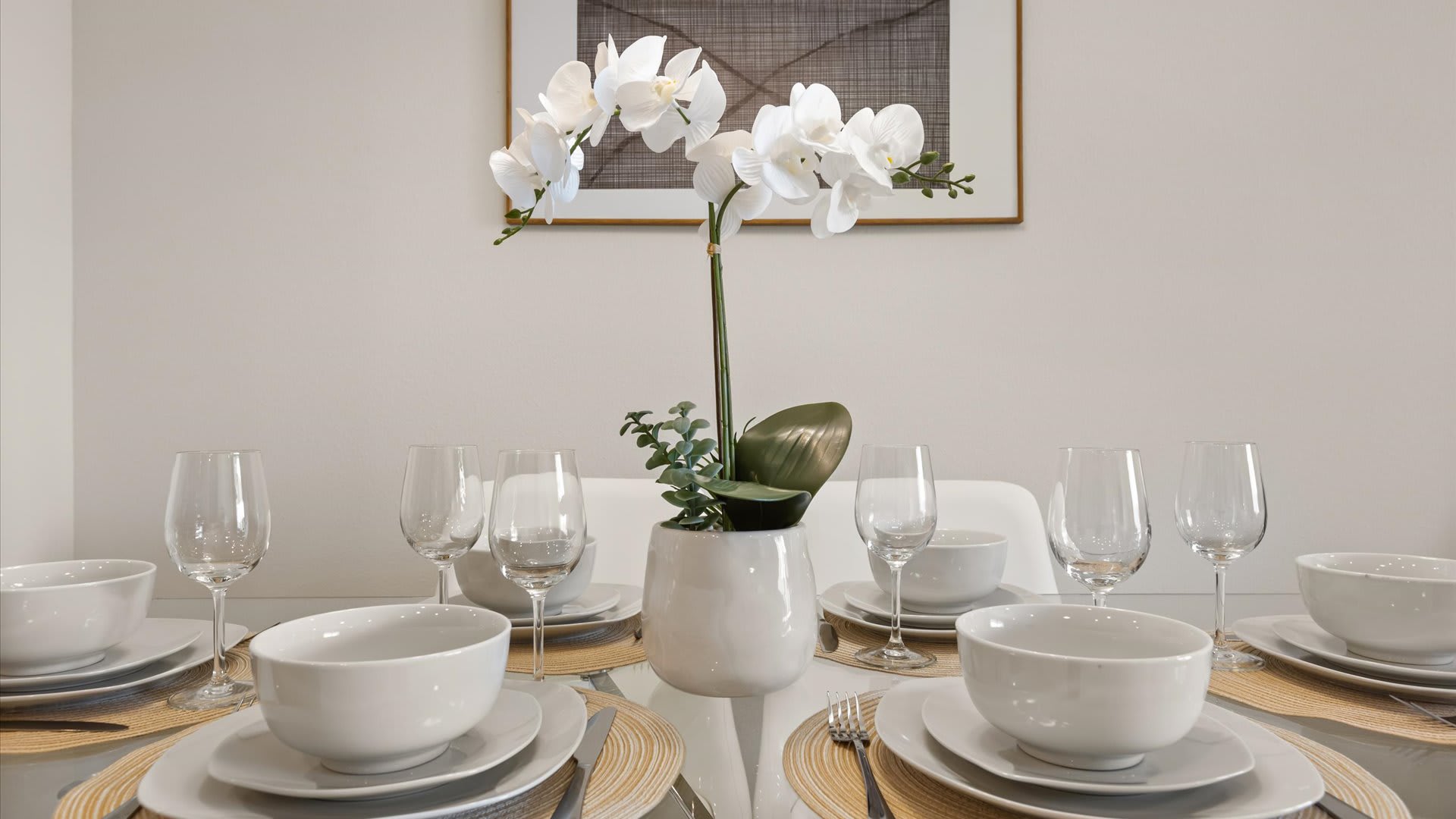 Elegant dining table set with white dinnerware, wine glasses, and a white orchid centerpiece, positioned against a light-colored wall with framed artwork.