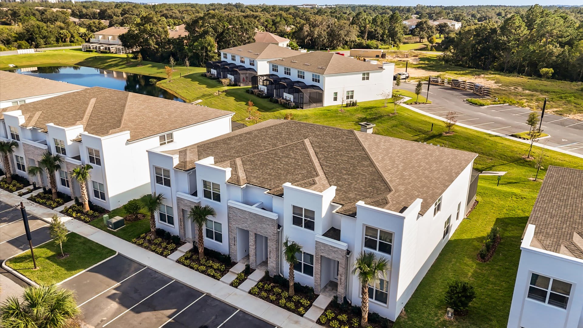 An aerial view of a modern townhouse community with white buildings, brown roofs, and stone accents, surrounded by green lawns, a parking lot, and a distant lake. The townhouses are arranged in rows with some under construction.