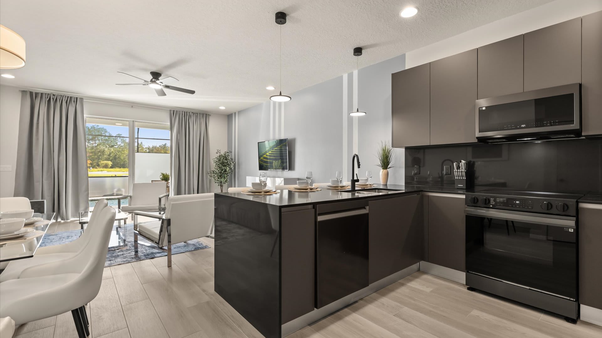 Modern kitchen with dark gray flat-panel cabinetry, black countertops, and stainless steel appliances, overlooking a dining area with a glass table and white chairs, and a living room with a view to the outdoors.
