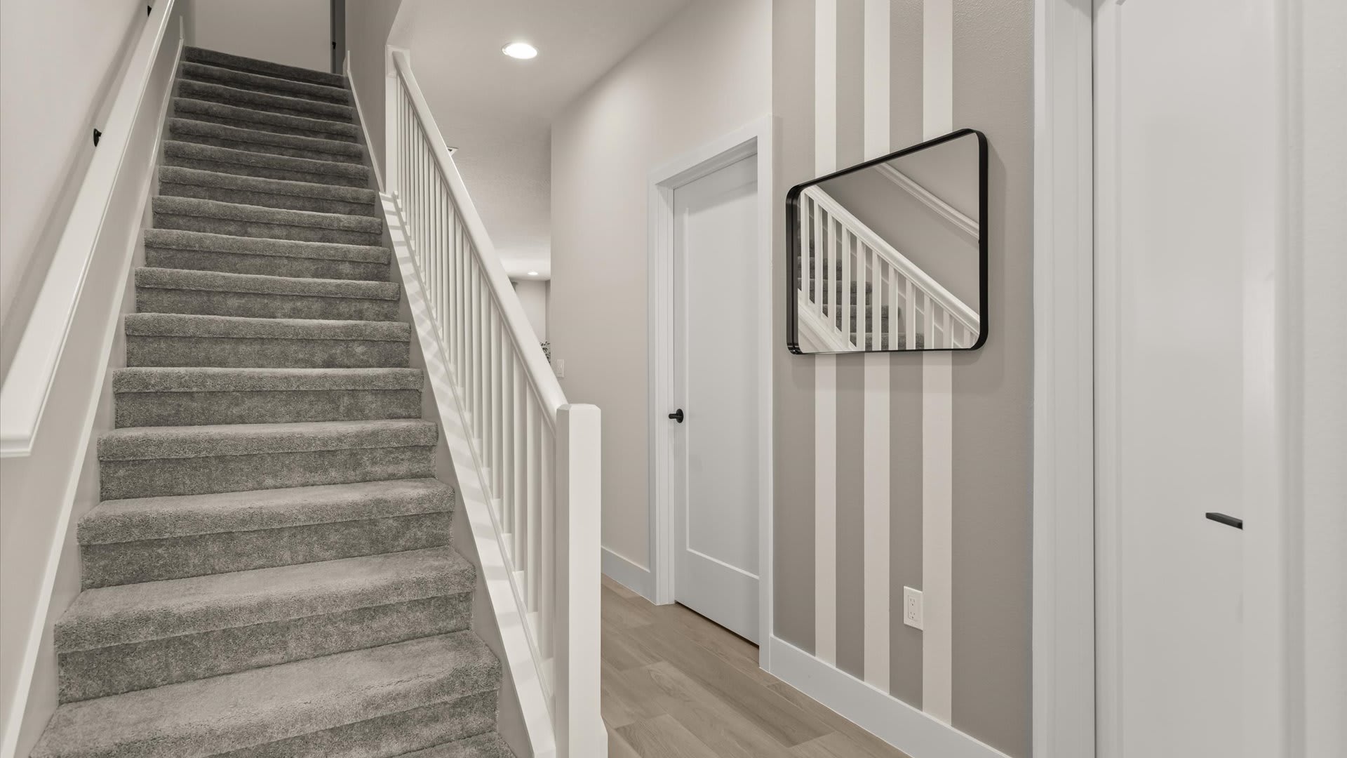 Hallway with light wood-look flooring leading to a carpeted staircase with white railings, featuring a gray accent wall with white stripes and a rectangular black-framed mirror.