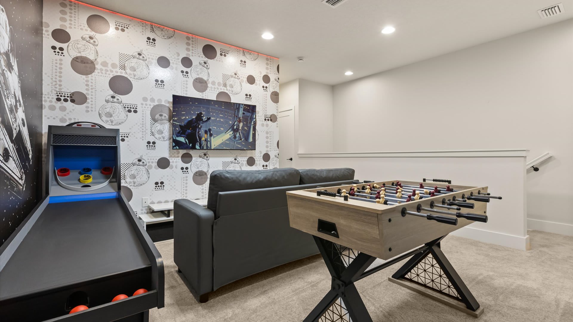 Star Wars-themed game room with a foosball table, a skee-ball style arcade game, a gray sofa, and a wall-mounted TV, featuring graphic wallpaper and red LED strip lighting.