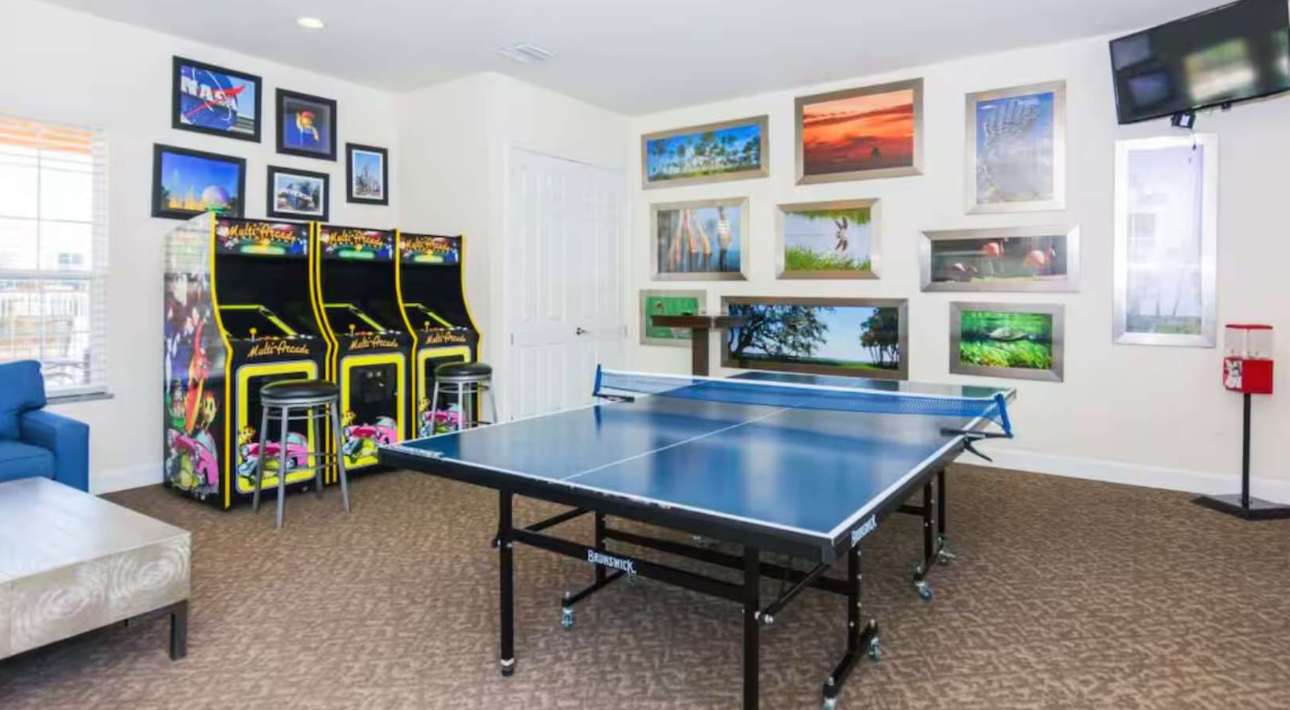 A vibrant game room featuring a blue ping-pong table in the center, two arcade machines with stools, and various framed artwork on the light-colored walls. A blue sofa and coffee table are also visible.
