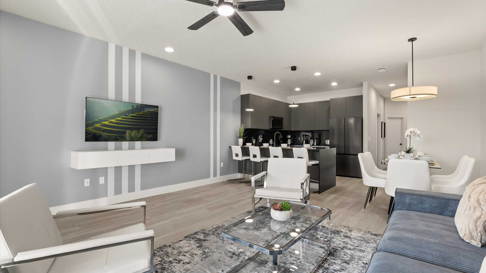 Open-concept living space with a wall-mounted TV on a gray accent wall with white stripes, two white armchairs, and a view into the modern kitchen and dining area with dark cabinetry and light flooring.