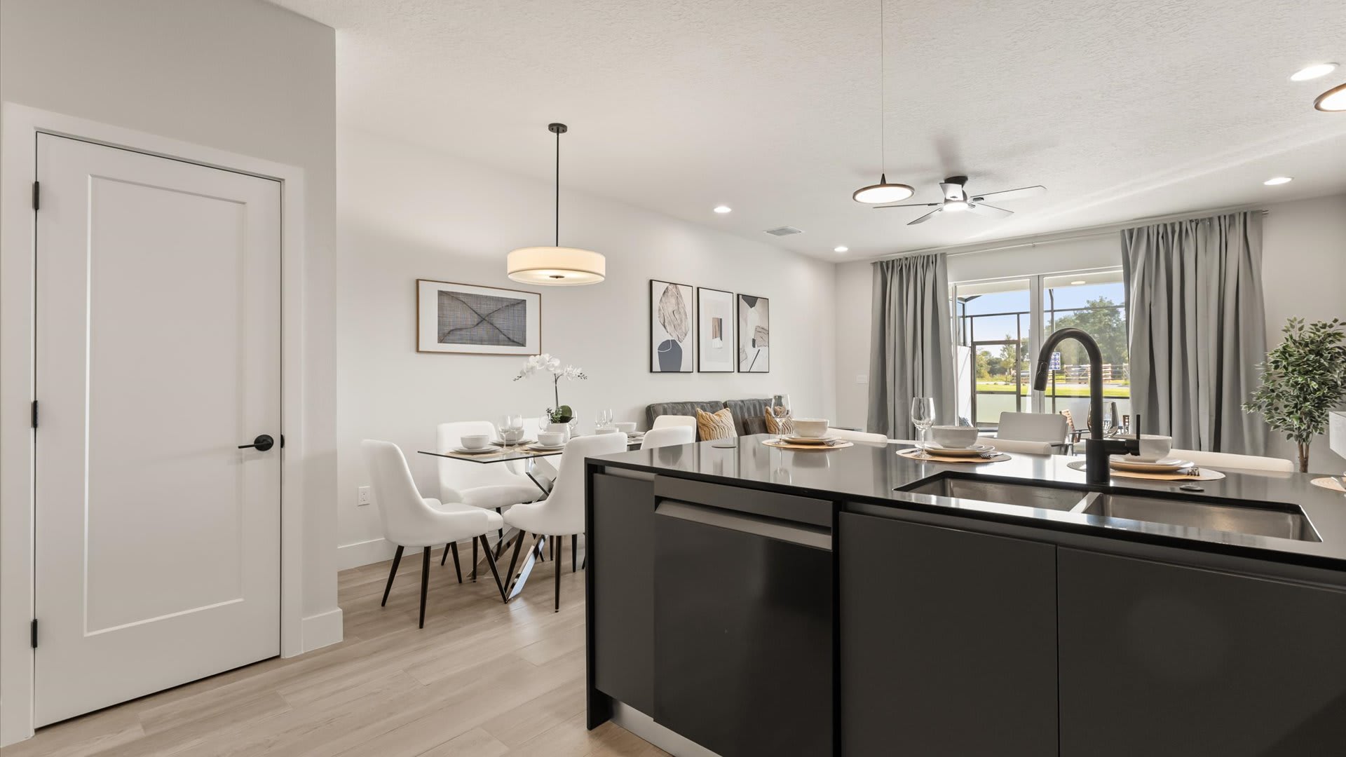 Bright dining area with a glass-top table and white chairs, adjacent to a modern kitchen with dark cabinetry and an island, and a view towards the living room and outdoor patio.