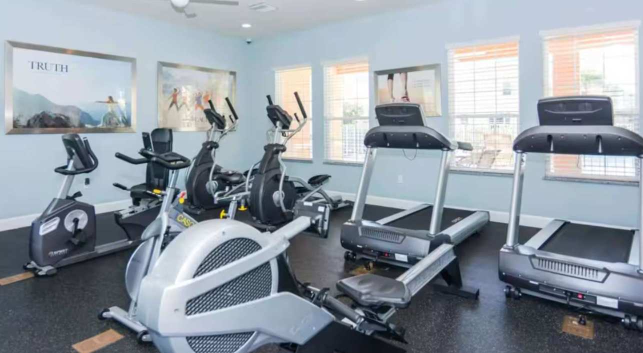 A well-equipped fitness room with various exercise machines including treadmills, ellipticals, and stationary bikes, set against light blue walls with large windows and framed artwork. The floor is dark rubber.