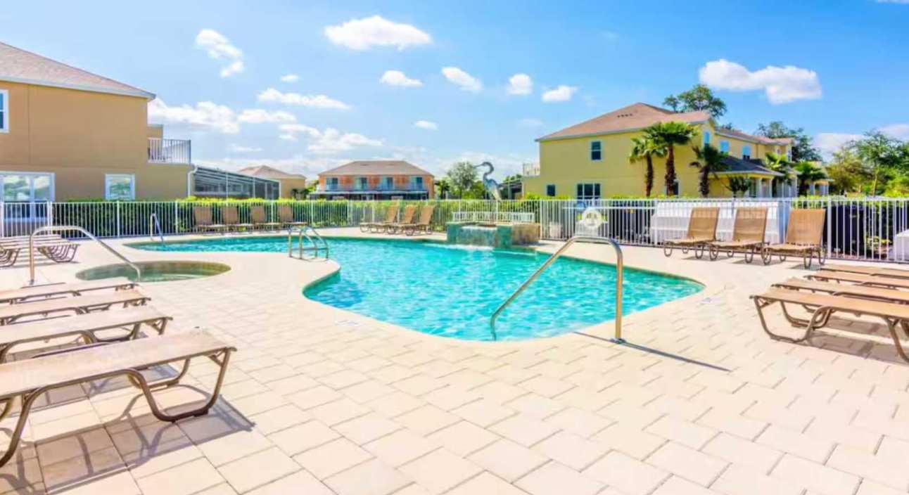 A communal outdoor swimming pool with a hot tub, surrounded by light-colored paving stones and several lounge chairs under a bright blue sky. Yellow-colored houses are visible in the background.