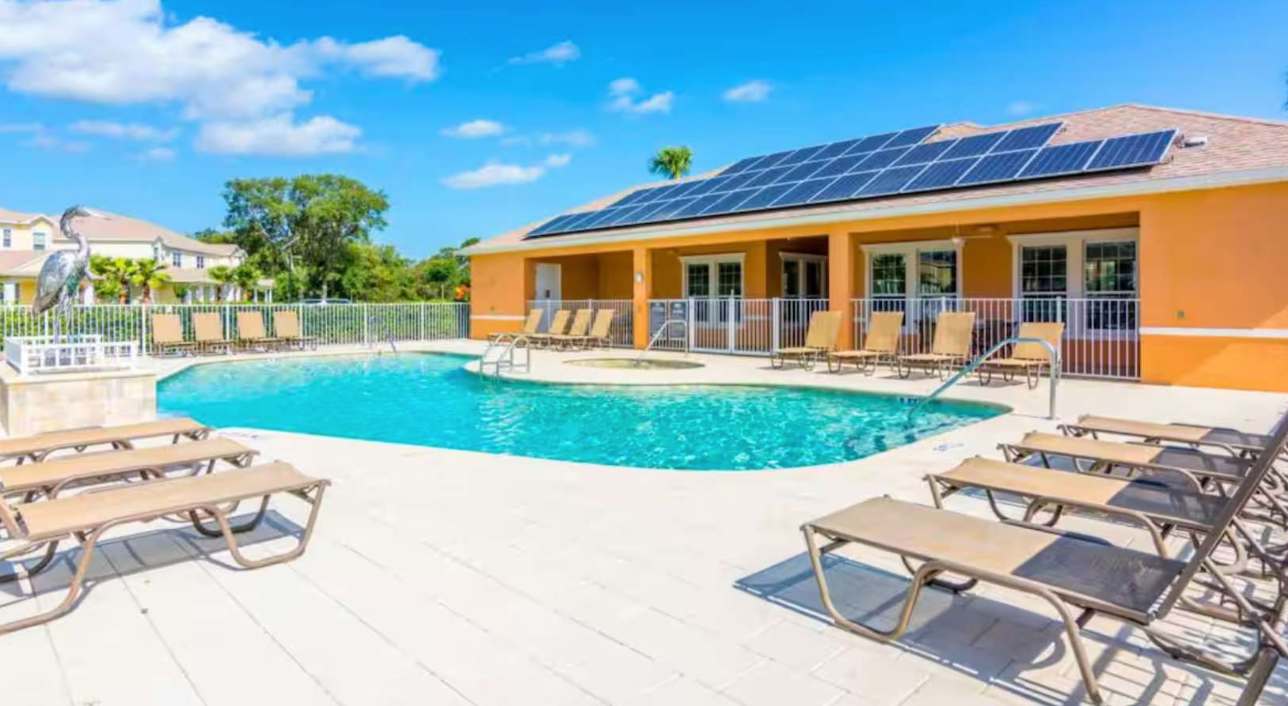 An outdoor communal pool area with a large swimming pool, a hot tub, and a clubhouse building with solar panels on its roof. Numerous lounge chairs are arranged on the surrounding paved deck.
