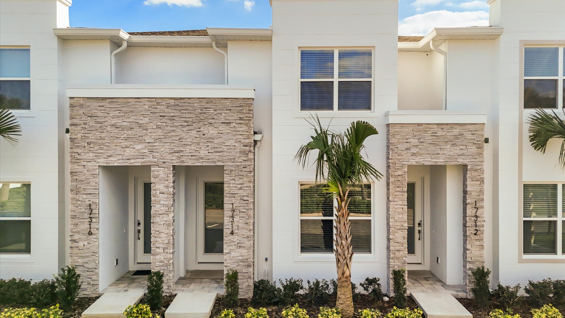 A modern white townhouse facade with light grey stone accents around the entrances, featuring multiple windows with blinds and a small palm tree in the foreground. House numbers '1734' and '1732' are visible on the doors.