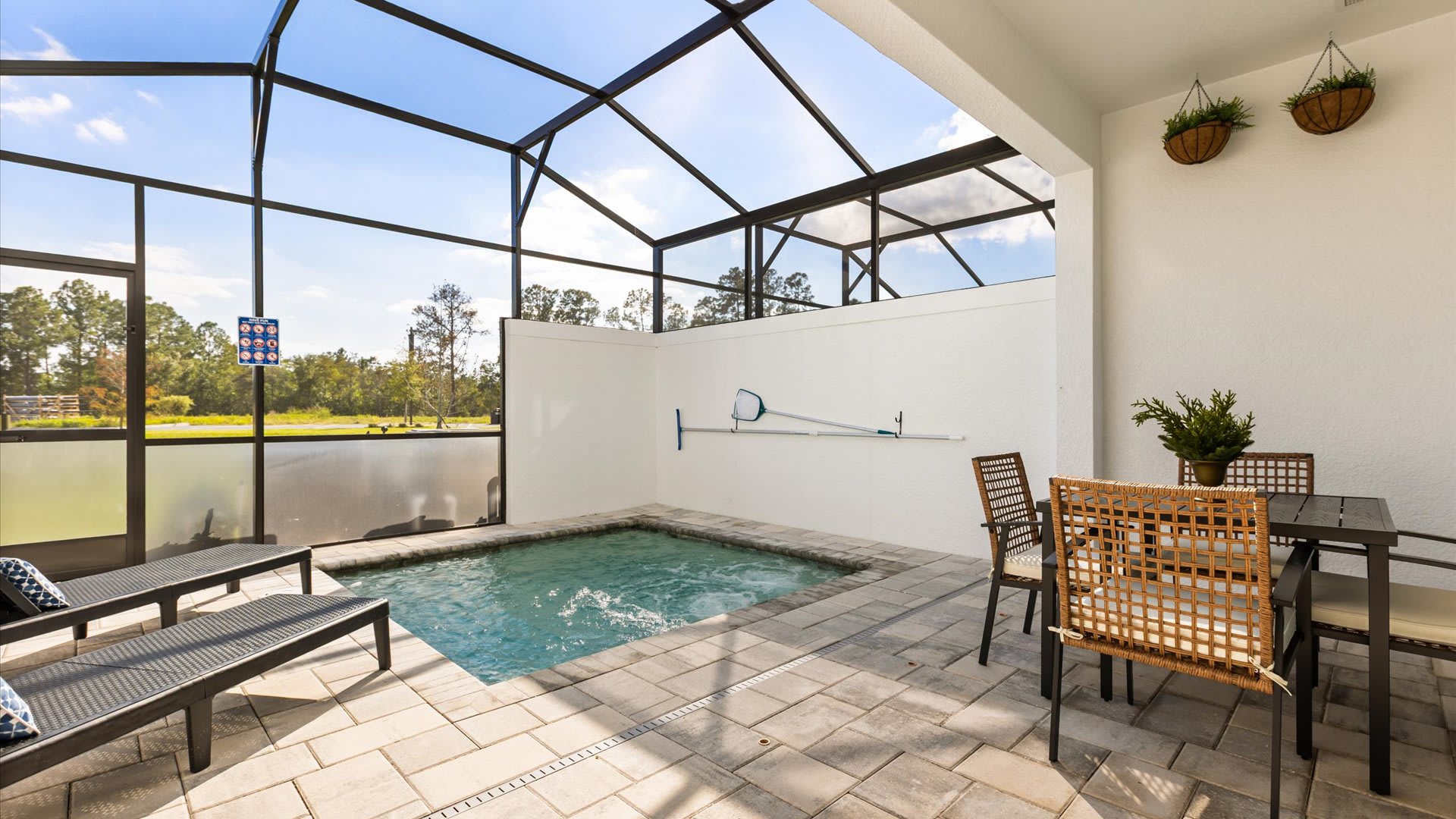 Screened-in outdoor patio featuring a bubbling hot tub, two lounge chairs with blue pillows, and a dining table with four chairs, all set on light-colored pavers under a clear sky.