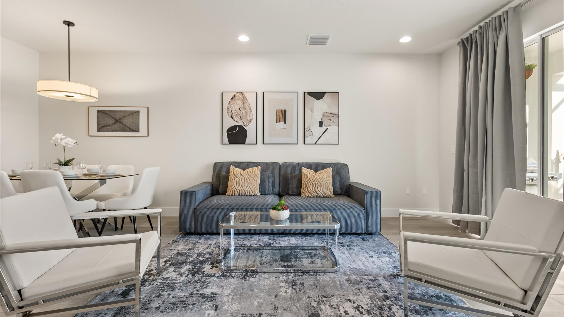 Bright living room with a gray sofa and two white armchairs on a blue and gray abstract rug, facing a glass coffee table, with a dining area visible in the background.