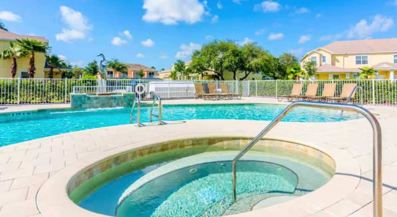 A close-up view of the circular hot tub integrated into the communal outdoor pool area, with clear blue water and stainless steel handrails. The main pool and lounge chairs are visible in the background.