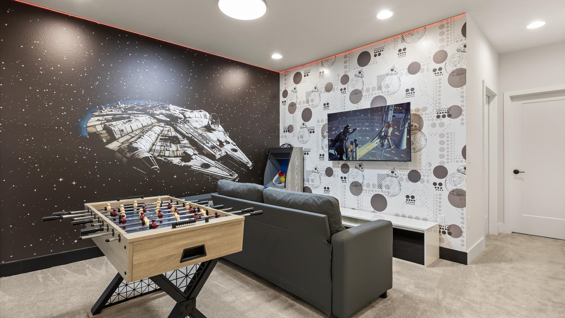 Immersive Star Wars-themed game room with a large Millennium Falcon mural on one wall, a foosball table, a gray sofa, and a wall-mounted TV, with patterned wallpaper and red LED lighting.