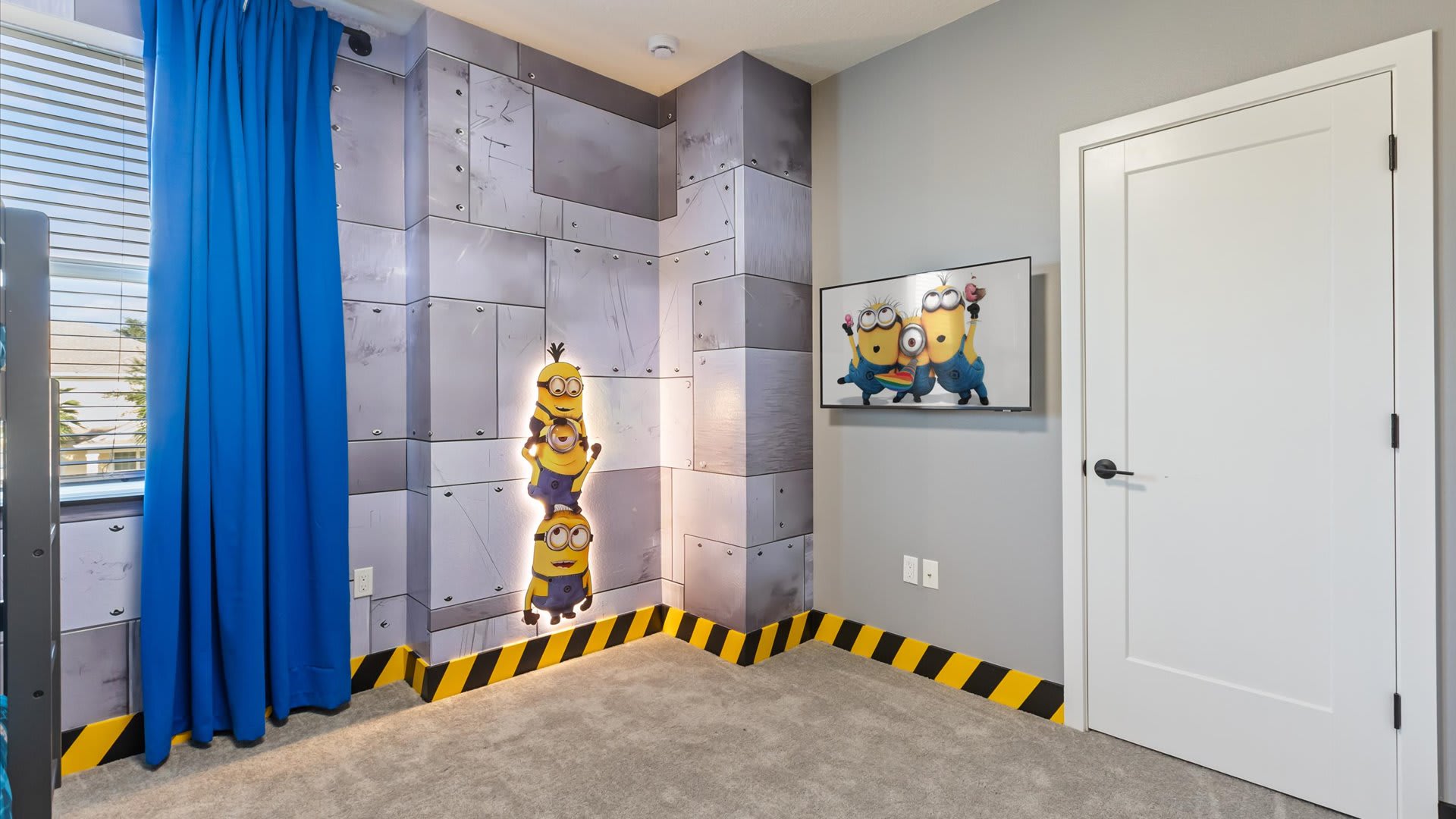 Corner of a Minions-themed kids' bedroom with industrial-style wall panels, a wall-mounted TV displaying Minions characters, and a white door. Yellow and black striped tape lines the base of the walls, adding to the theme.