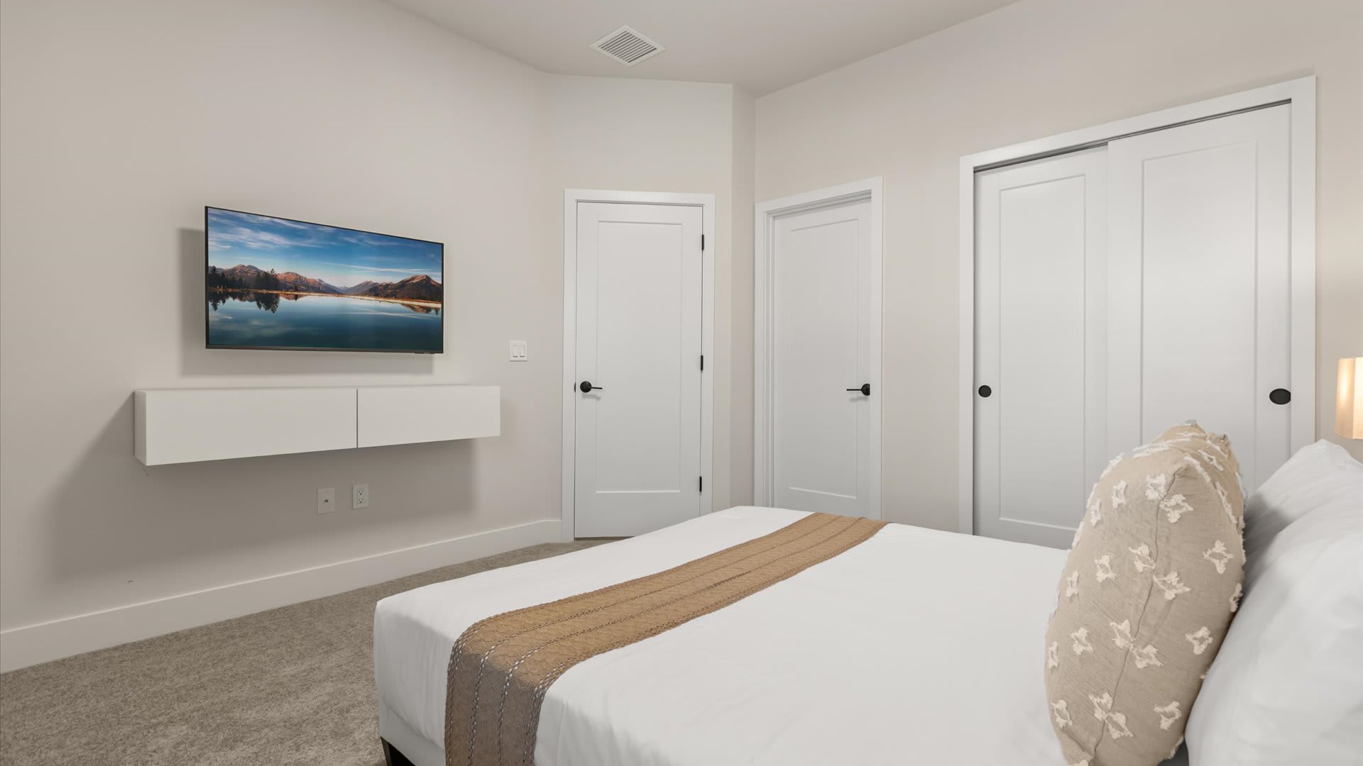 Neutral-toned bedroom with a wall-mounted flat-screen TV above a floating white console, and two white doors, one leading to a closet with sliding doors. A bed with neutral bedding is partially visible in the foreground.