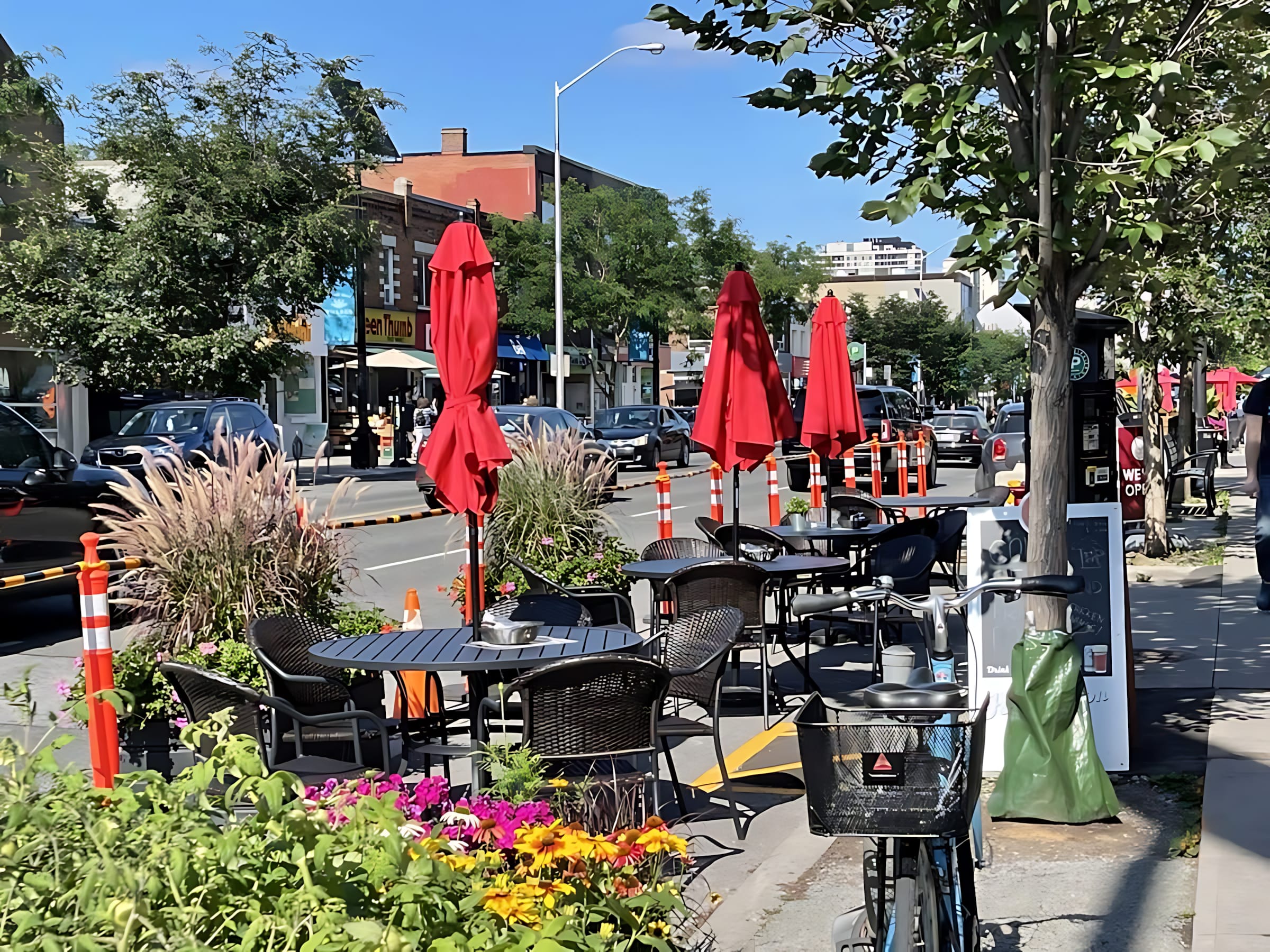 A lively street scene featuring an outdoor patio dining area with black wicker chairs and tables, shaded by red umbrellas, alongside a street with parked cars and a bicycle in the foreground.