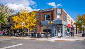 An exterior view of a brick commercial building on a sunny day, featuring a cafe named 'Baka Gallery Cafe' with outdoor seating under a yellow tree, and people walking on the sidewalk.
