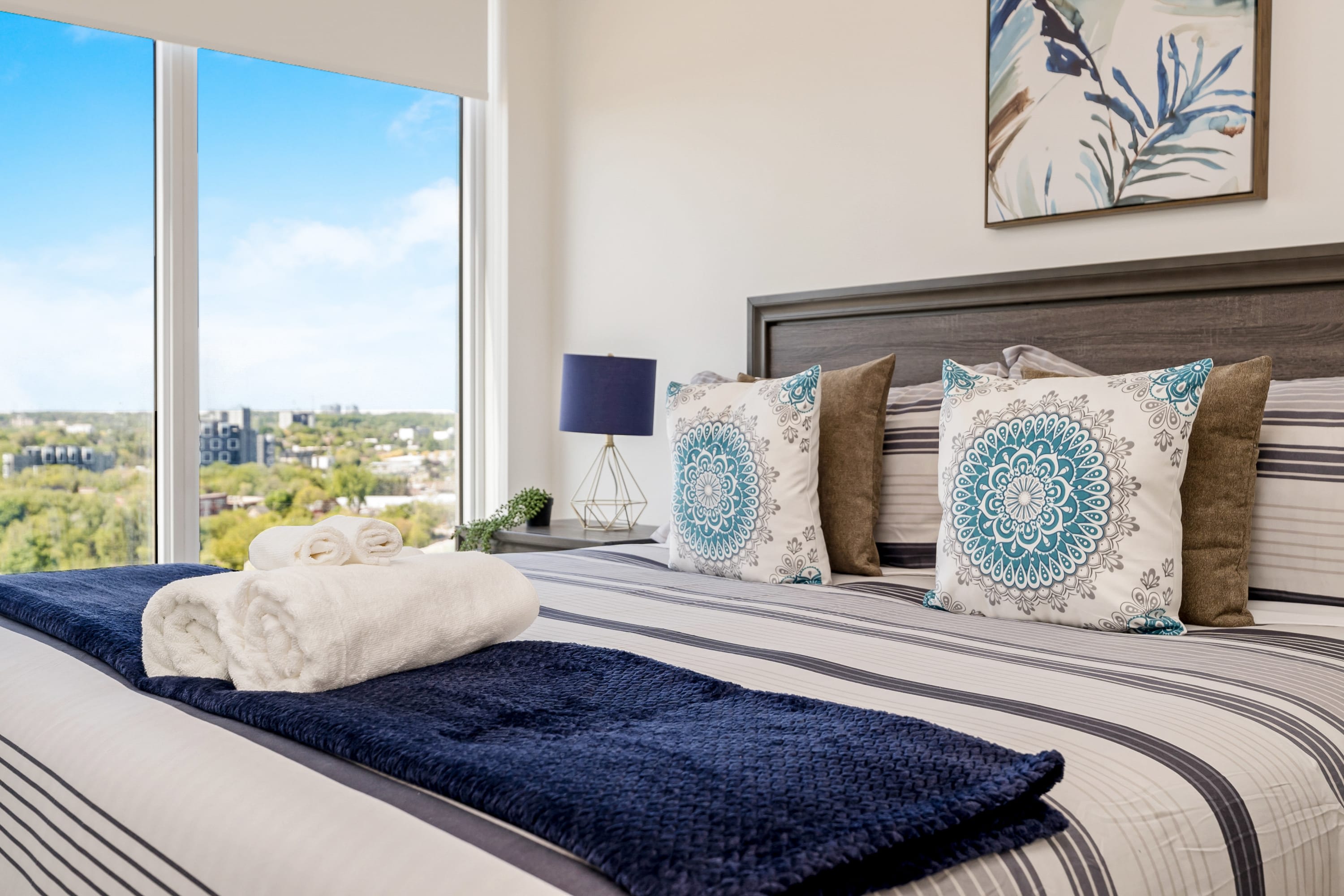 A close-up of the bedroom shows the bed with striped bedding, decorative pillows, and a blue throw, with a nightstand and lamp visible. A large window offers a panoramic view of a city skyline under a bright blue sky.