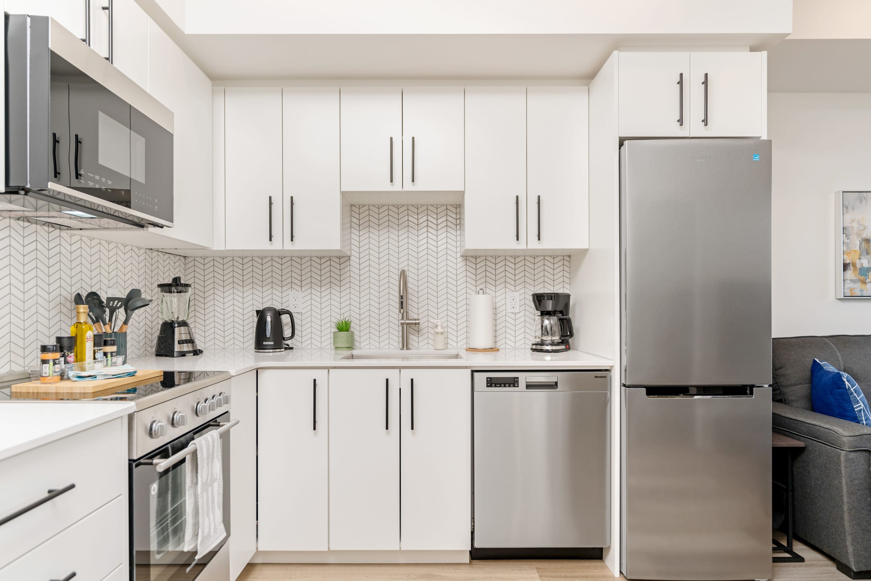 Close-up view of the modern white kitchen with stainless steel appliances, including a refrigerator, oven, microwave, and dishwasher, set against a white herringbone tile backsplash.