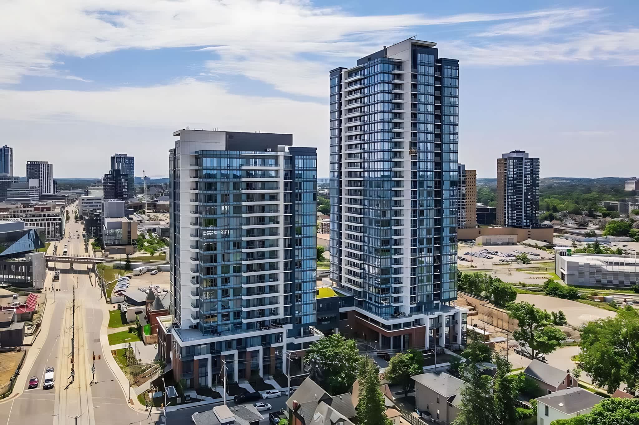 An aerial view showcases two modern high-rise condominium buildings with glass facades and balconies, situated in an urban setting with surrounding streets, smaller buildings, and green spaces under a partly cloudy sky.