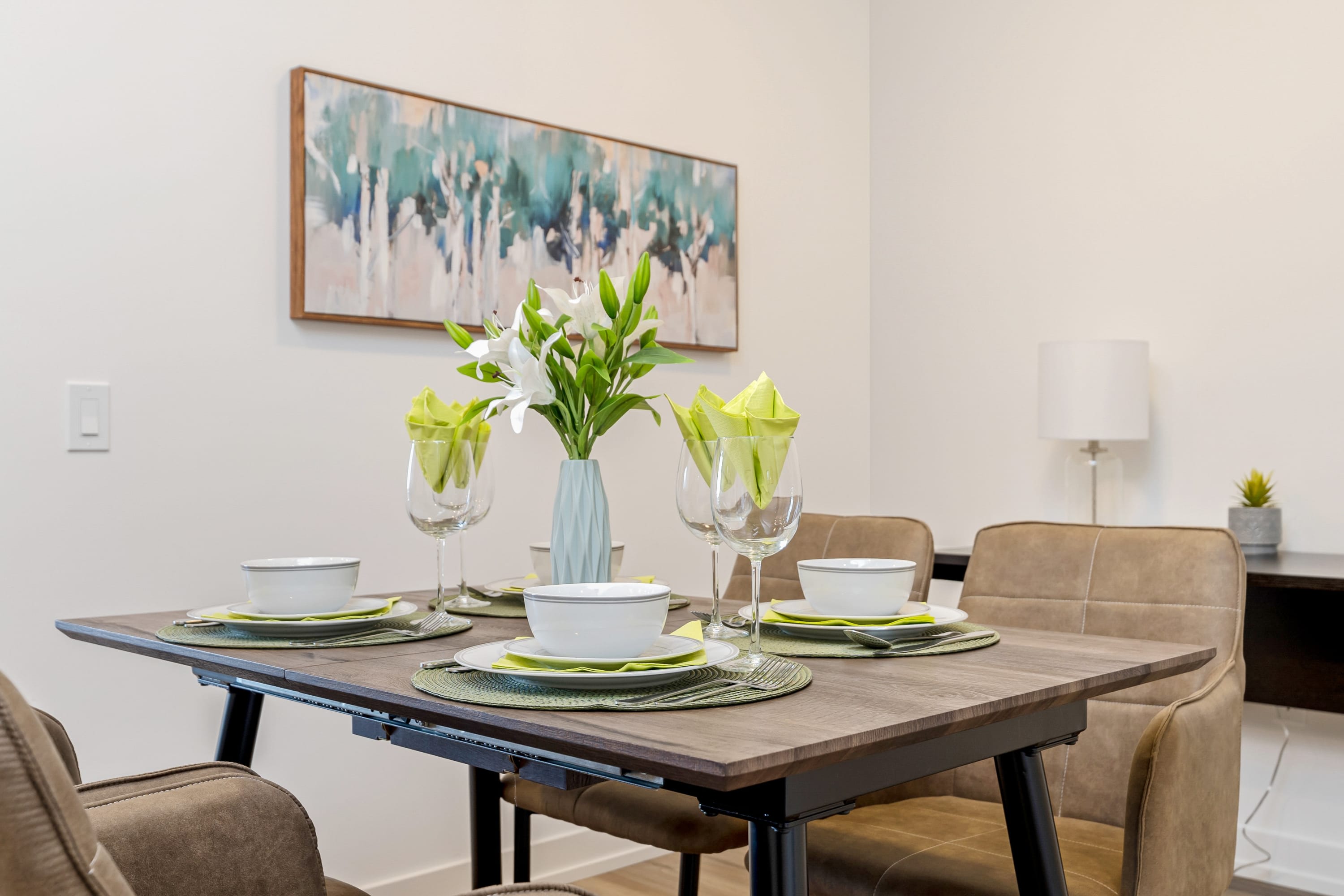 A modern dining area features a dark wood table set for four with light green placemats and white dishware, complemented by a light blue vase with white lilies and a large abstract painting on the white wall.
